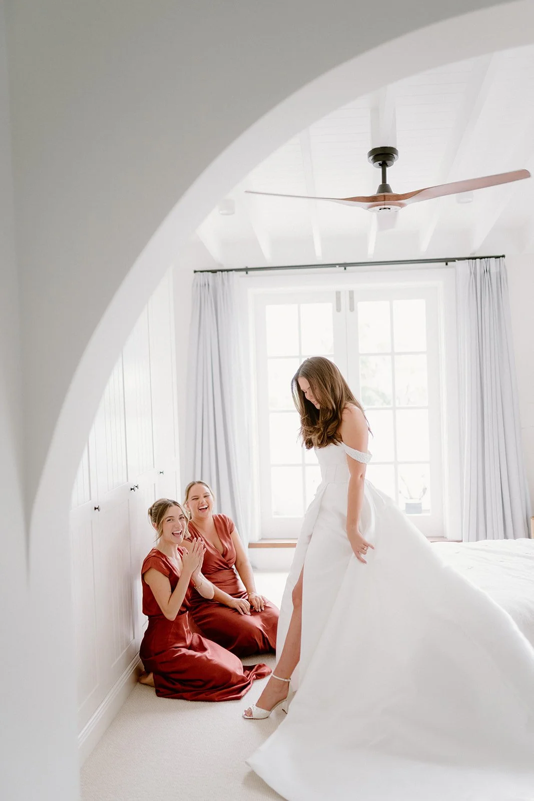 A bride in a white wedding dress standing in a bright room with two bridesmaids in red dresses kneeling on the floor, all happily laughing and smiling.