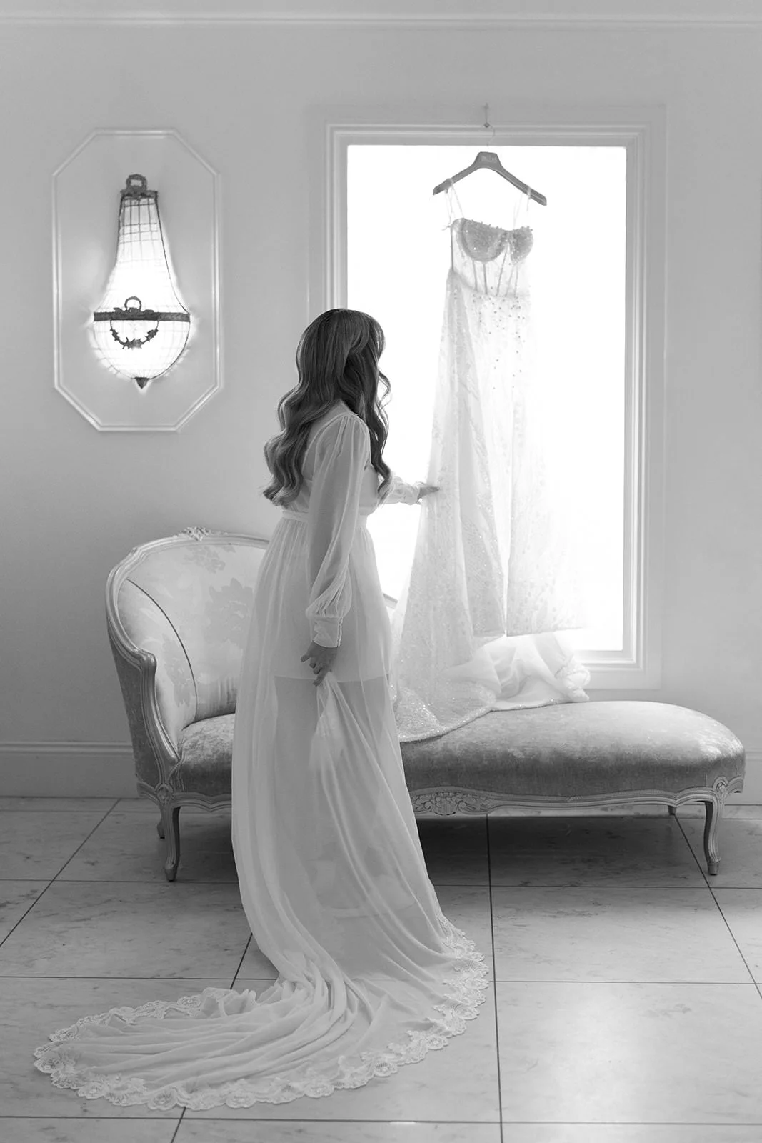 A woman in a wedding dress gazing at a hanging wedding gown in front of a large window, in a room with vintage sofa and wall sconce lighting.