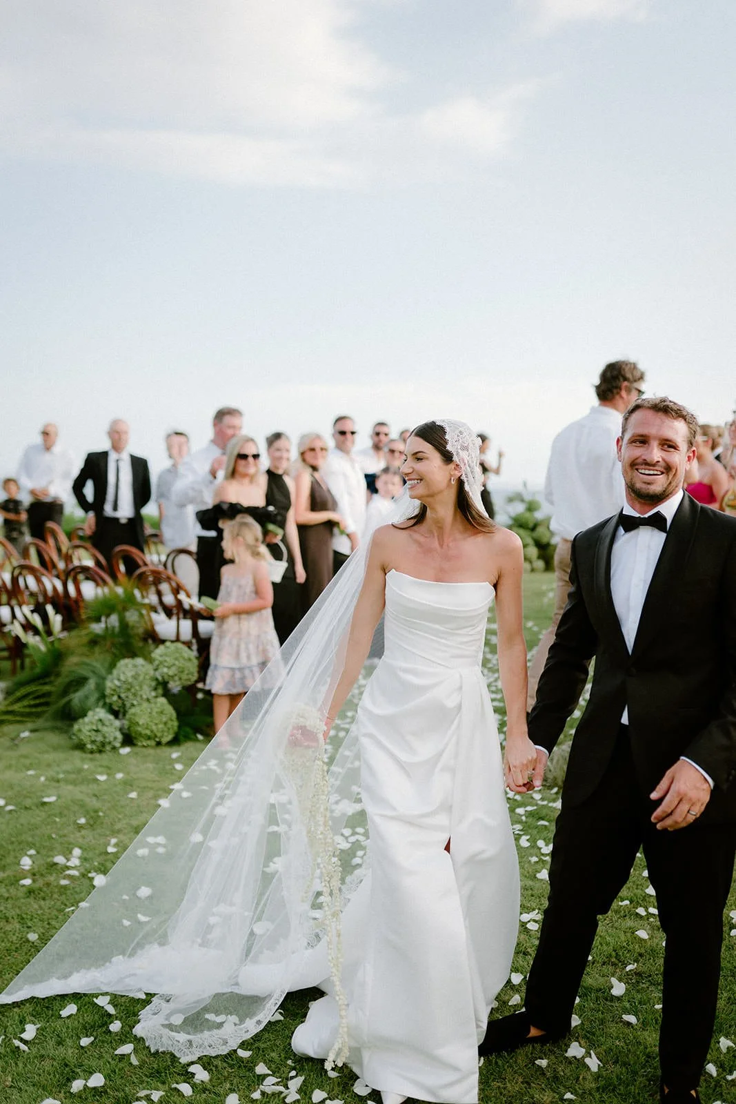 A newlywed couple walking hand in hand outdoors during a wedding ceremony, with guests in the background.