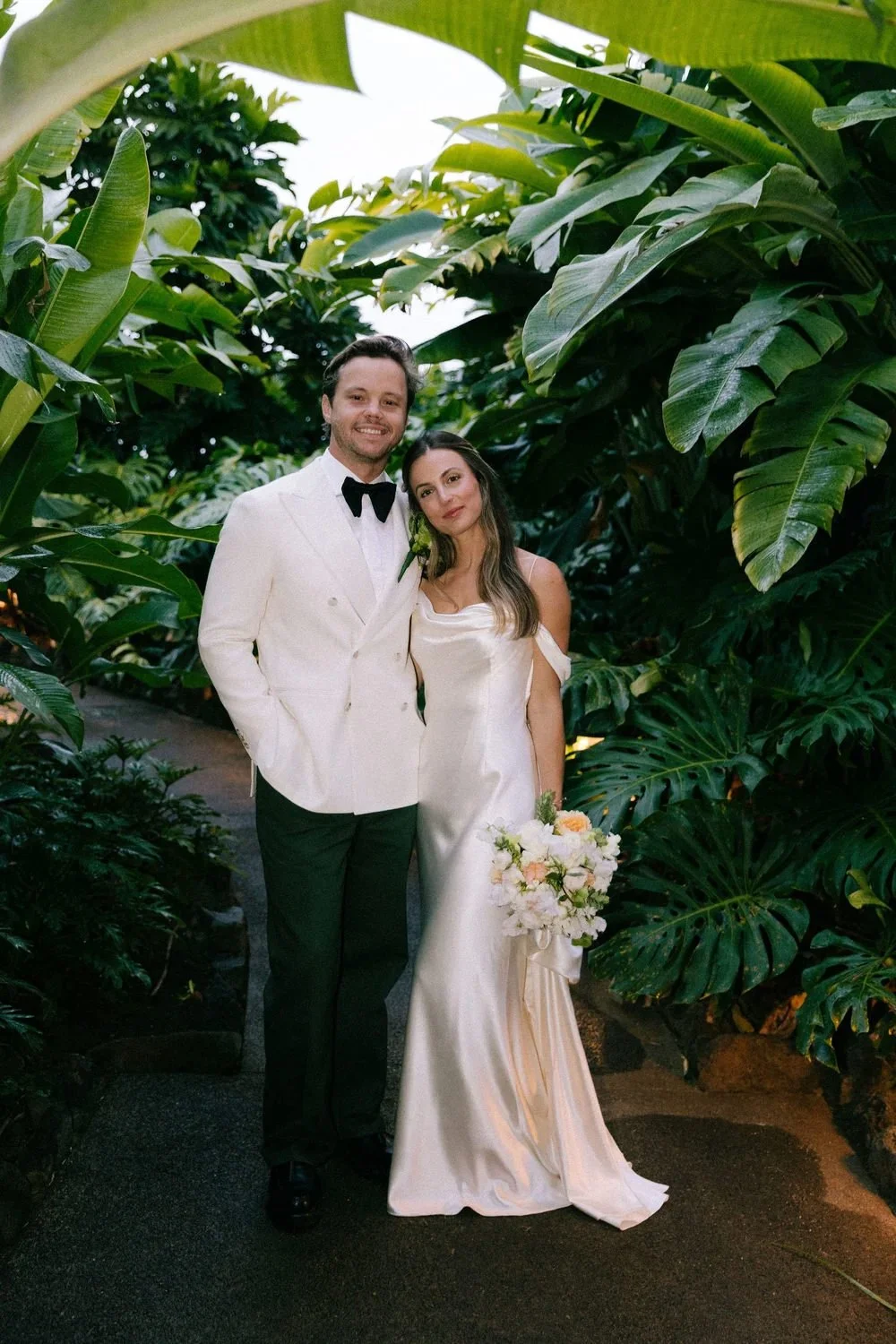 A newlywed couple standing close together in a lush tropical garden, with the groom wearing a white tuxedo and black bow tie, and the bride in a satin off-shoulder wedding gown holding a bouquet of white and pink flowers.