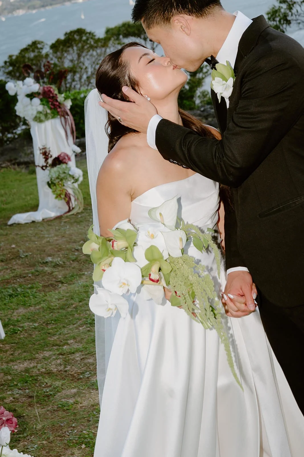 Bride and groom sharing a kiss at their outdoor wedding, with the bride in a white gown holding a cascading bouquet of white orchids and greenery, and the groom in a black tuxedo with a white boutonniere.