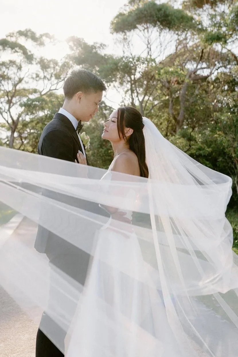 A bride and groom standing close together outdoors on a sunny day, gazing into each other's eyes. The bride wears a white wedding dress and veil, while the groom is in a black tuxedo. The background features trees and a clear sky.
