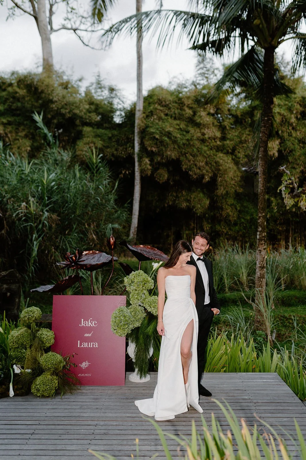 A bride and groom standing together on a wooden platform outdoors during a wedding ceremony, with lush green trees and plants in the background. The bride is wearing a strapless white gown with a high slit, and the groom is wearing a black tuxedo wit