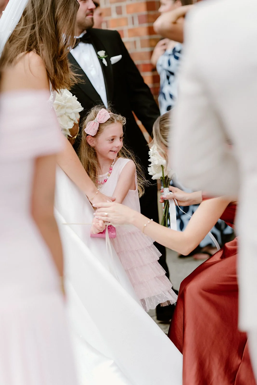 A young girl in a pink dress with a pink bow headband, smiling, holding hands with a woman while receiving a flower from another girl in a maroon dress at a wedding ceremony.
