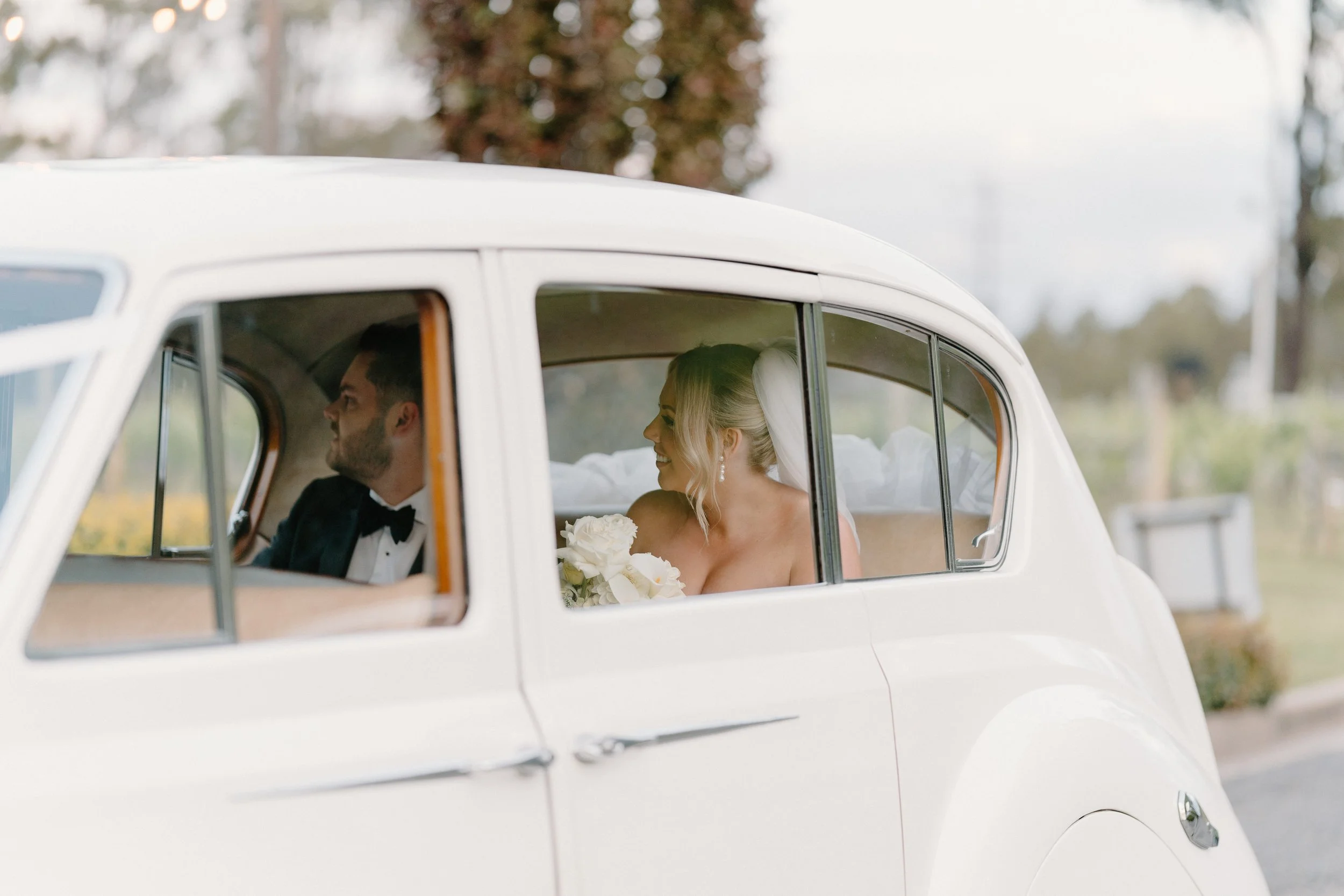 Bride and groom sitting in the back of a vintage white car on their wedding day, with the bride holding a bouquet of white flowers, smiling, in an outdoor setting with trees and cloudy sky in the background.