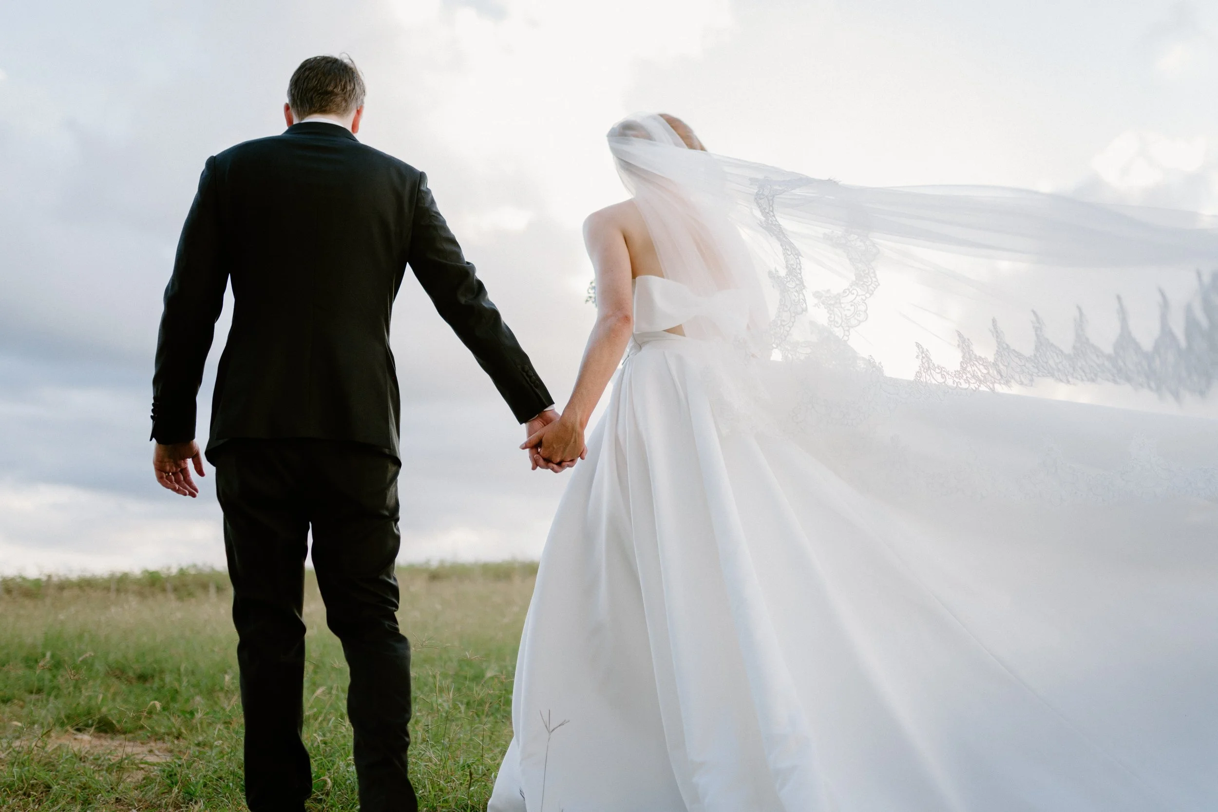 A bride and groom holding hands, walking outdoors on a grassy field with a cloudy sky in the background. Peterson House Wedding.