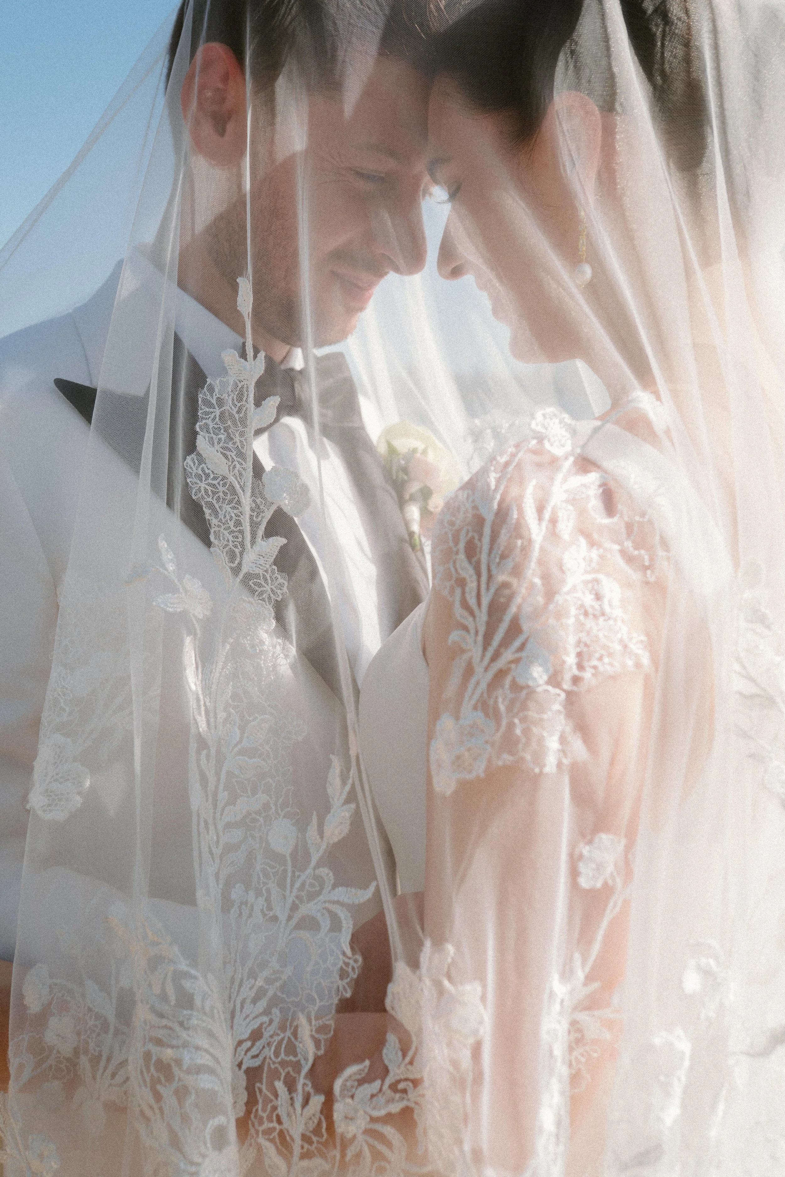 A bride and groom under a wedding veil, smiling and touching foreheads, outdoors during the day.