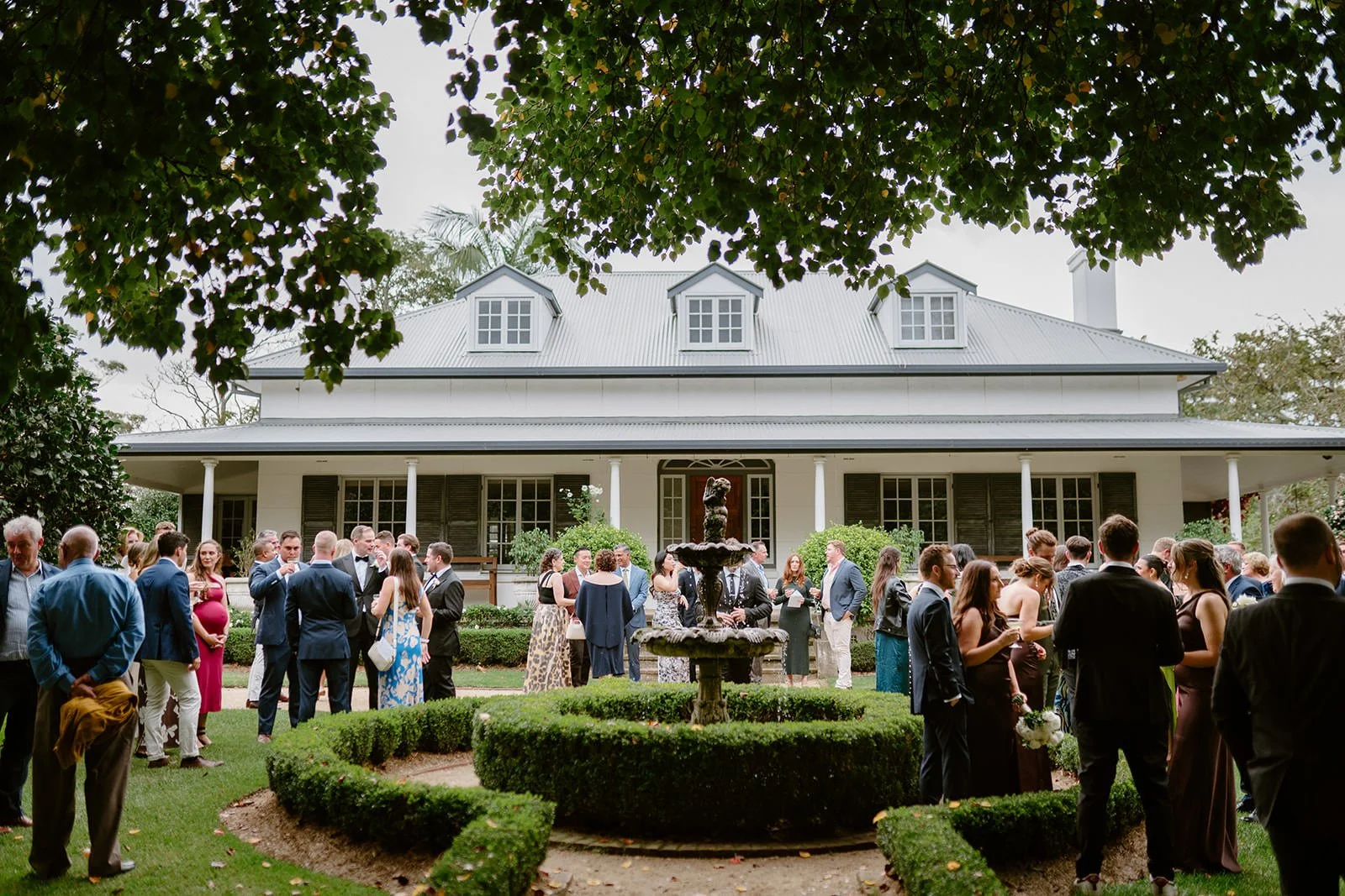 People gathered outdoors in front of a large white house with a lawn and fountain, likely at a social event or wedding reception.