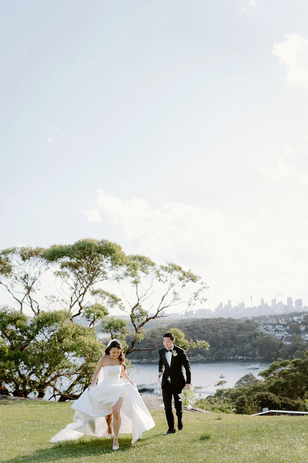 A bride and groom outdoors near a body of water, with trees and a city skyline in the background, during a wedding celebration.