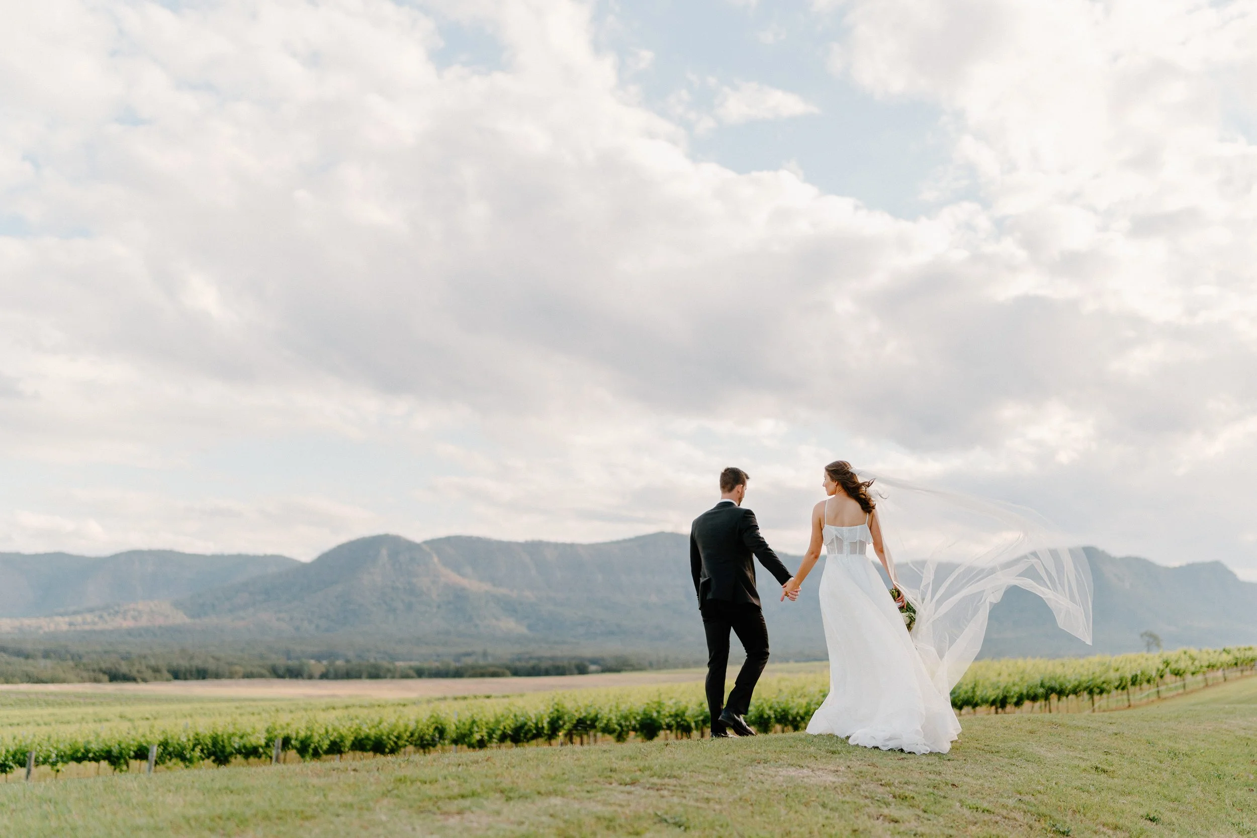 A bride and groom holding hands and walking on a grassy field with a scenic mountain range in the background and a cloudy sky above.