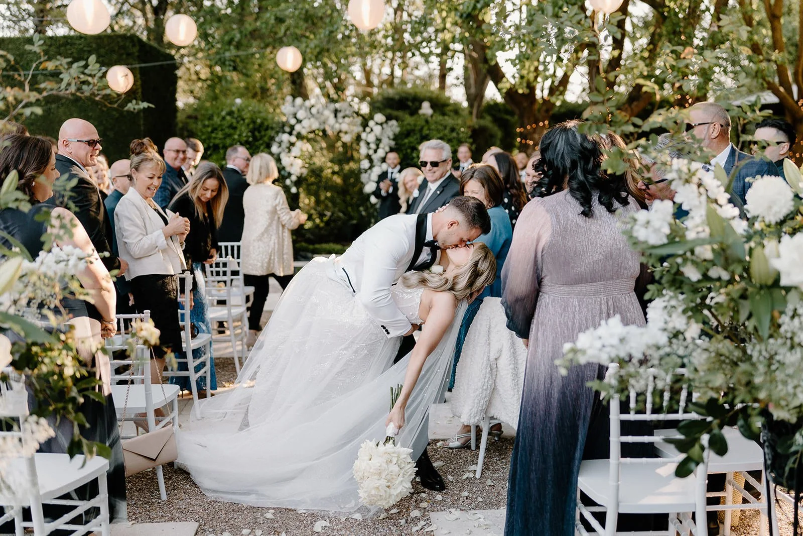 A newlywed couple shares a kiss during their outdoor wedding ceremony, with friends and family surrounding them, decorated with string lights, flowers, and greenery.