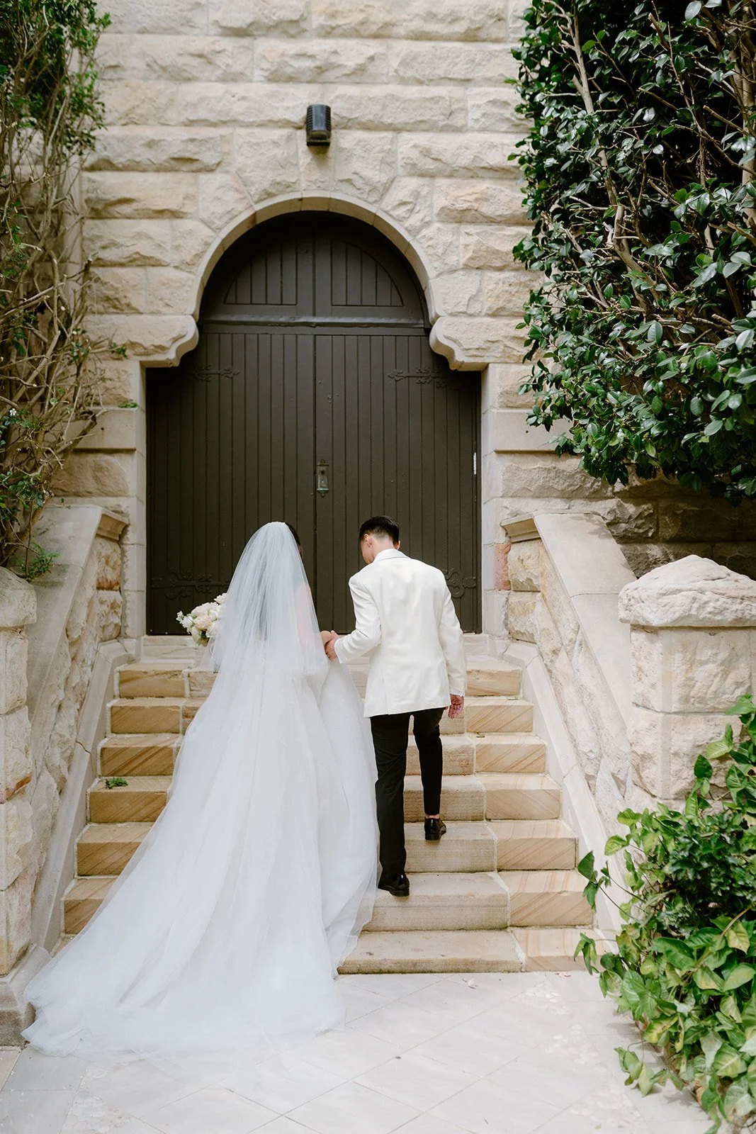 A bride and groom ascending stone stairs outside a building with a large black door, surrounded by greenery.