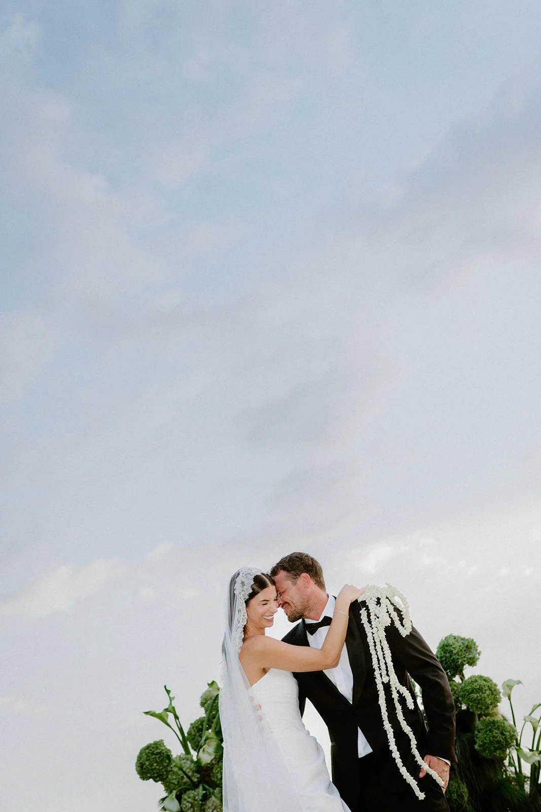 A bride and groom sharing a joyful moment outdoors on their wedding day, with the bride wearing a white wedding dress and veil, and the groom in a black tuxedo, decorated with white flowers.