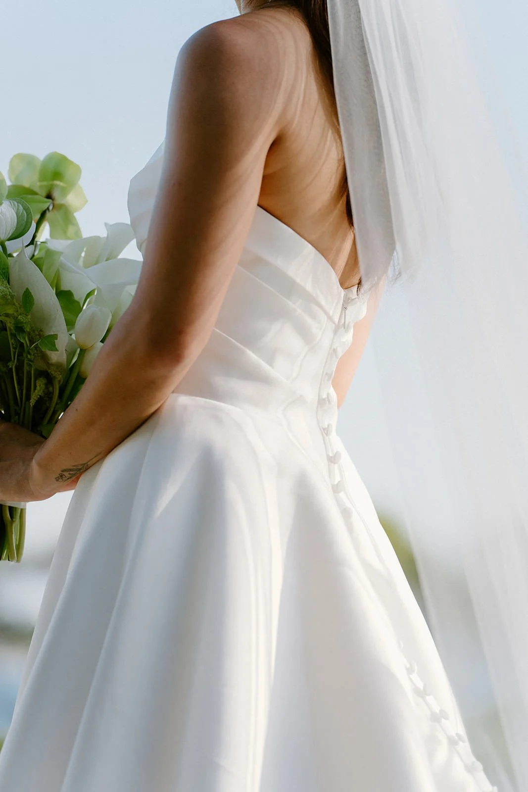 Close-up of a woman in a white wedding dress holding a bouquet of white flowers, standing near a window with sheer white curtains.
