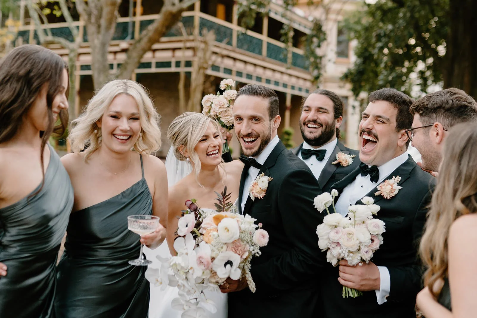 A group of wedding guests celebrating outdoors, with smiling men and women in formal attire, holding bouquets and drinks.