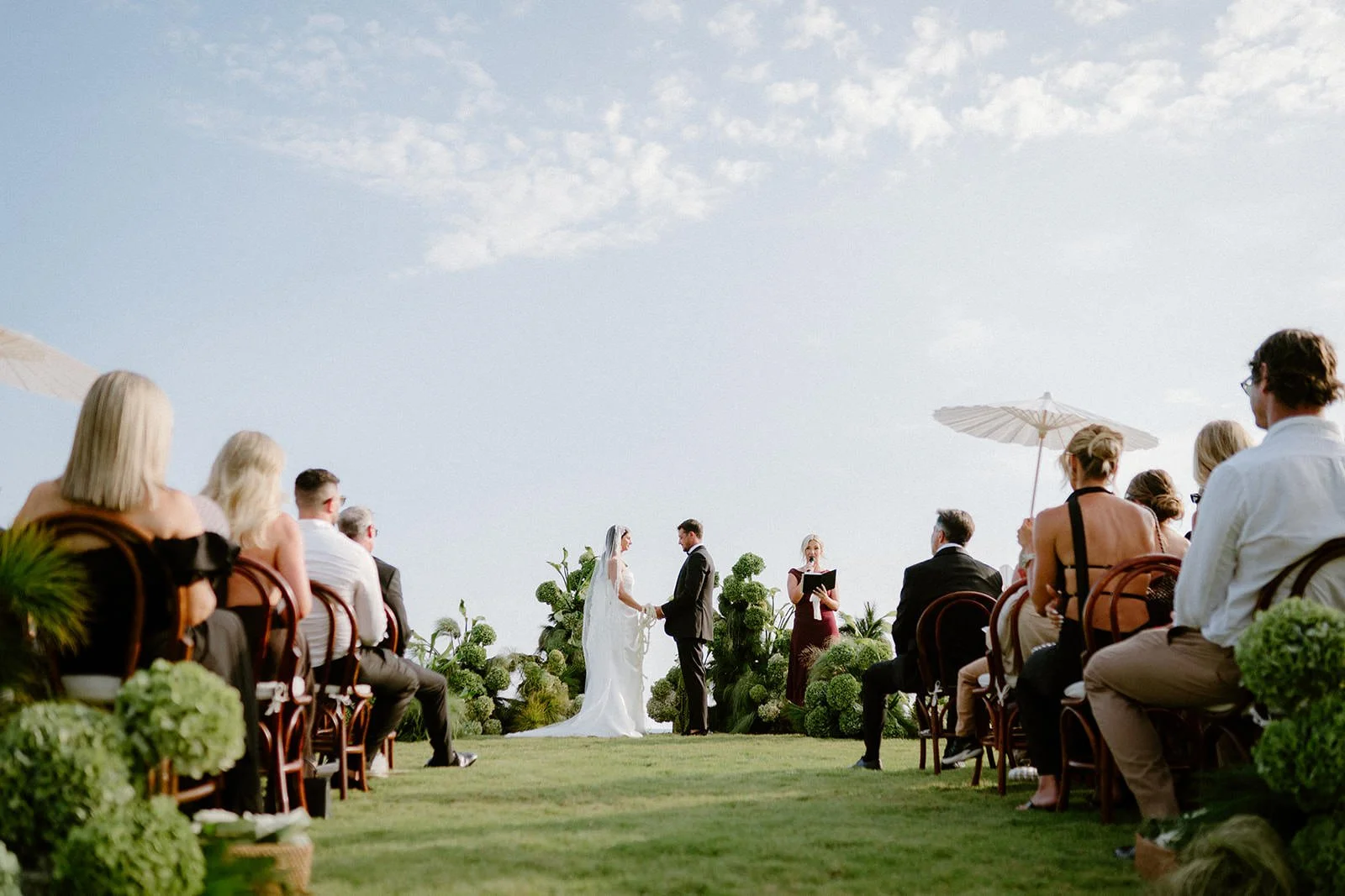 Bride and groom holding hands at an outdoor wedding ceremony on a grassy lawn, surrounded by guests seated on wooden chairs, with large green plants and white umbrellas in the background under a partly cloudy sky.