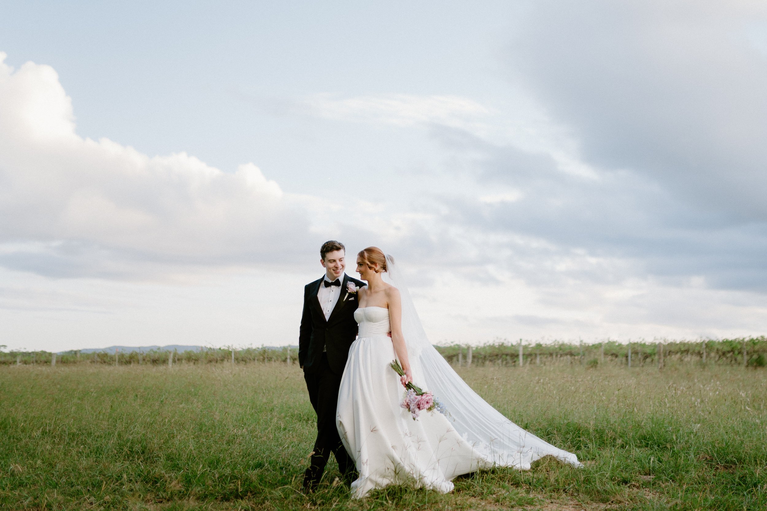 A bride and groom walking in a grassy field on a cloudy day. The bride wears a strapless white gown with a long train and veil, holding a bouquet of pink flowers. The groom wears a black tuxedo with a bow tie, smiling at the bride.
