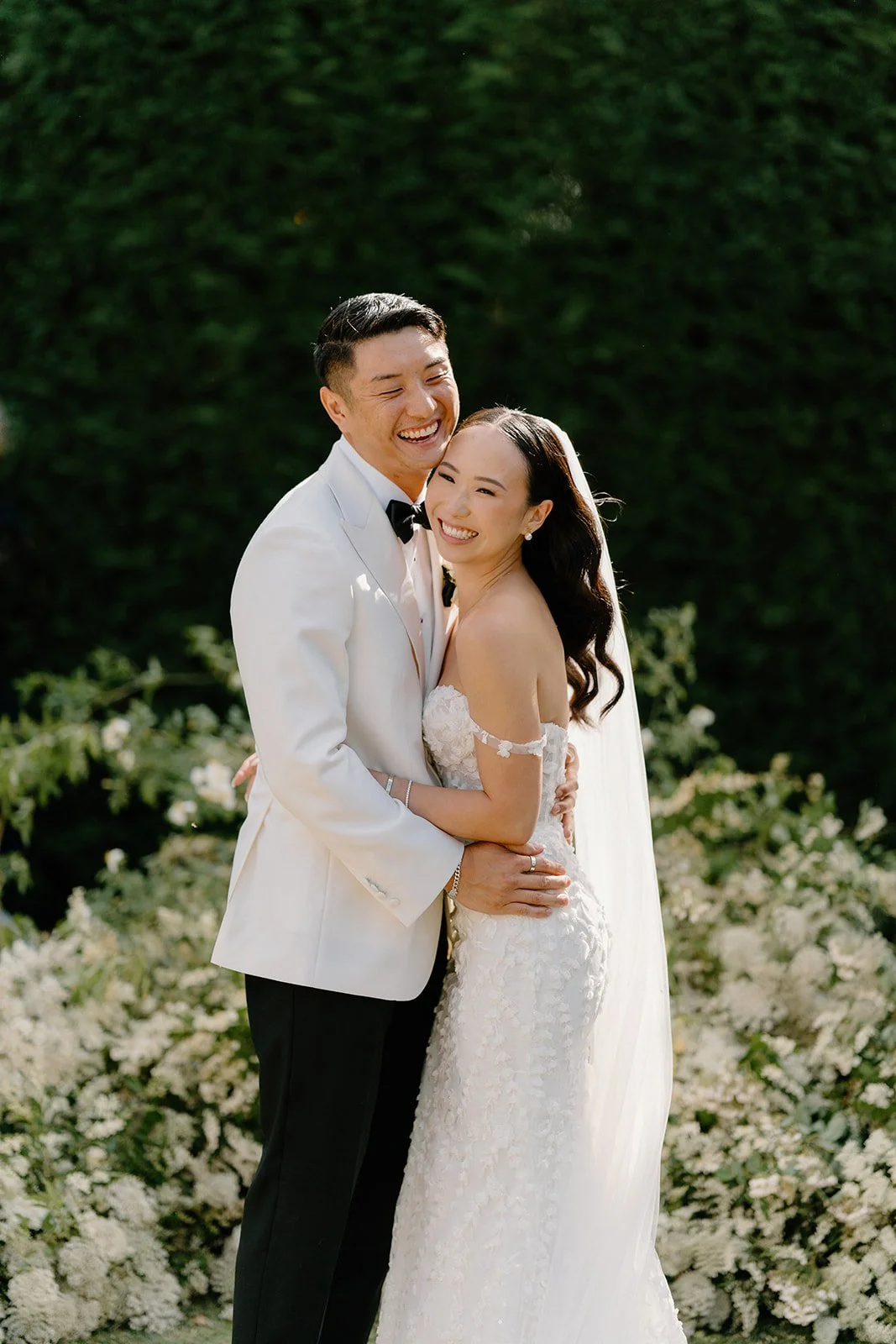 Couple dressed in wedding attire, the man in a white suit and black bow tie, the woman in a white lace wedding dress, hugging and smiling outdoors with greenery and white flowers in the background.