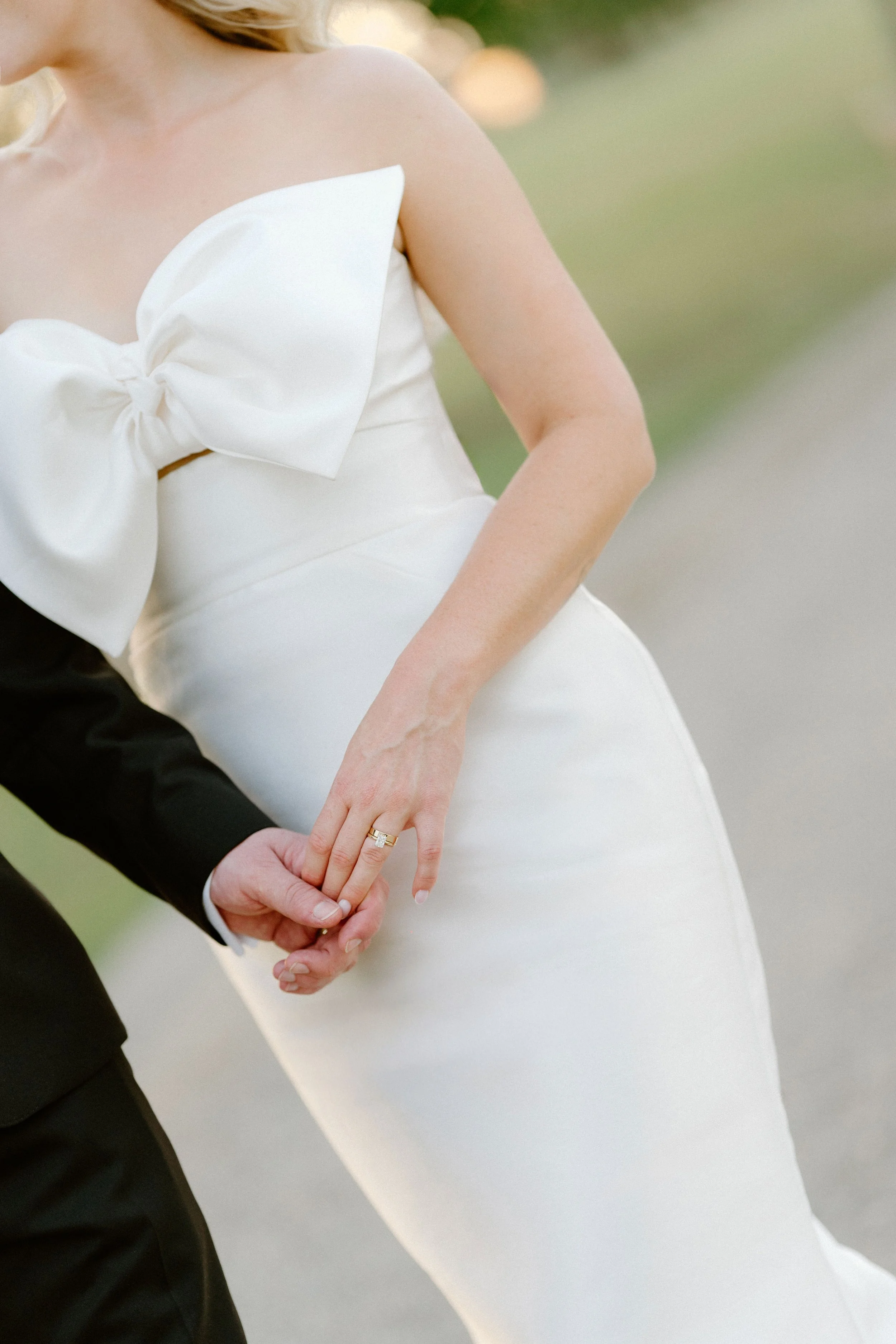 Close-up of a bride in a white wedding dress with a large bow on the chest, holding hands with a groom dressed in a black suit, on an outdoor path.