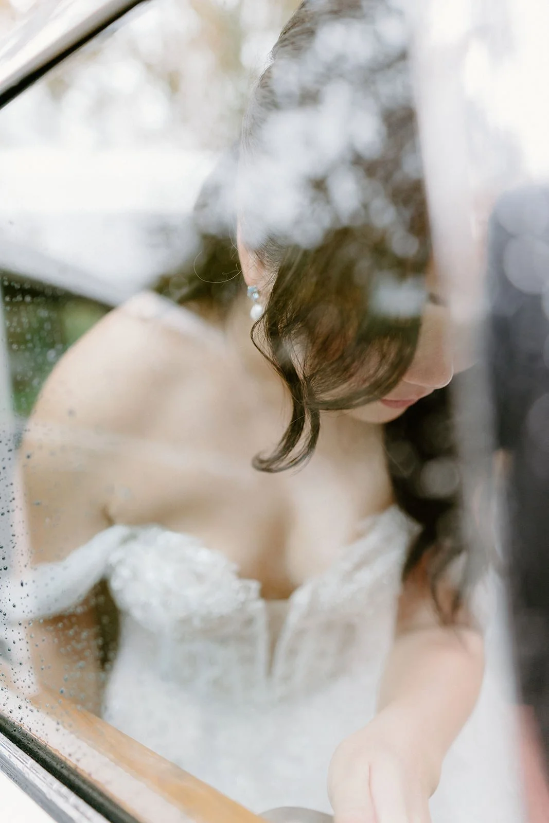A woman in a wedding dress viewed through a rainy window, looking down with wet hair and decorative earrings.