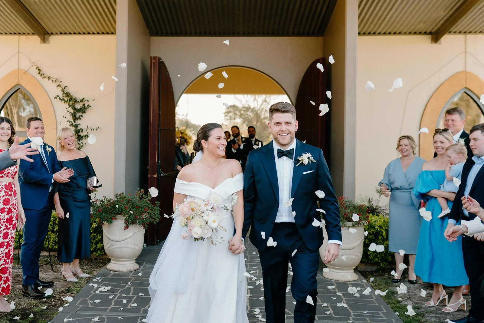 A newlywed couple holding hands and smiling as they walk out of a building, surrounded by friends and family celebrating with flower petals.