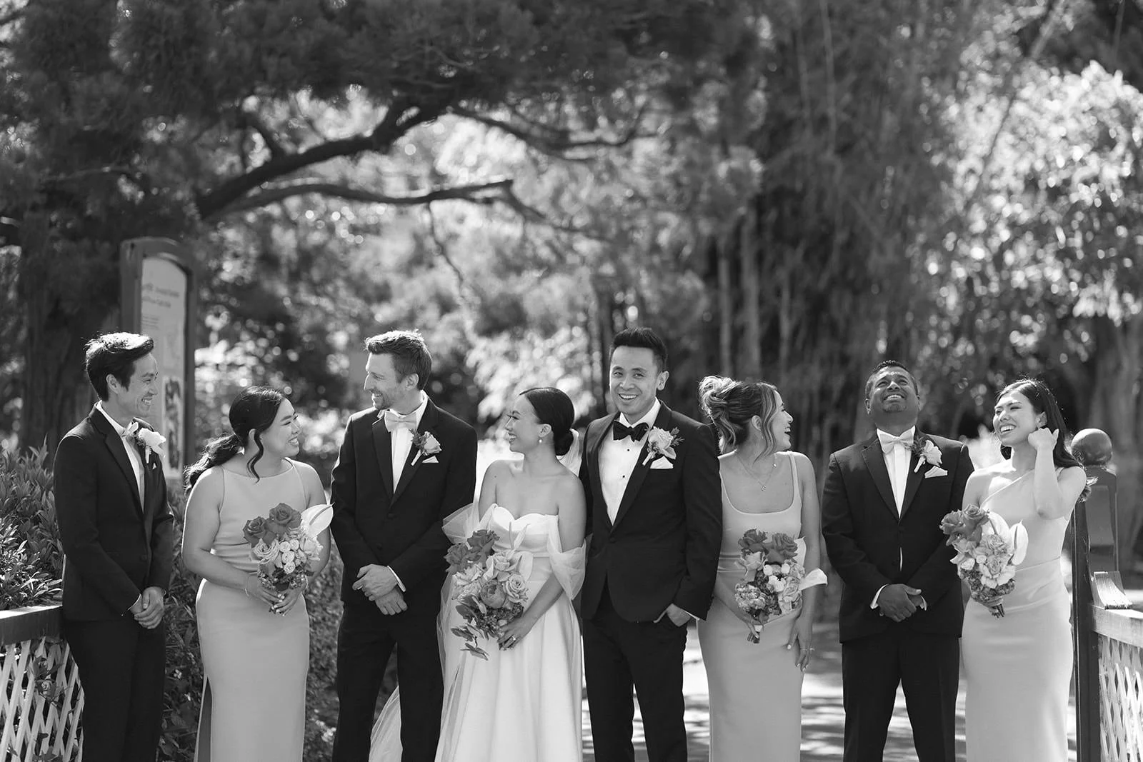 A wedding party outside, with eight people dressed in formal attire, smiling and standing close together in a line, surrounded by trees.