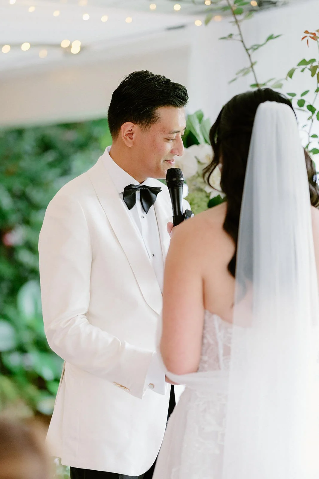 A groom in a white tuxedo and black bow tie is holding a microphone, standing in front of a bride with a veil, during a wedding ceremony with greenery in the background.