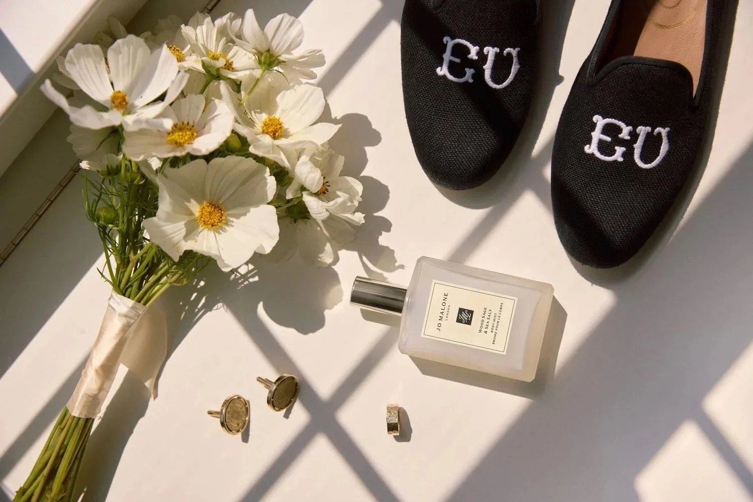 A bouquet of white flowers, a pair of black shoes with 'EU' embroidered on them, a bottle of Jo Malone perfume, a pair of gold cufflinks, a ring, and a small box of jewelry on a white surface with shadows from a window.