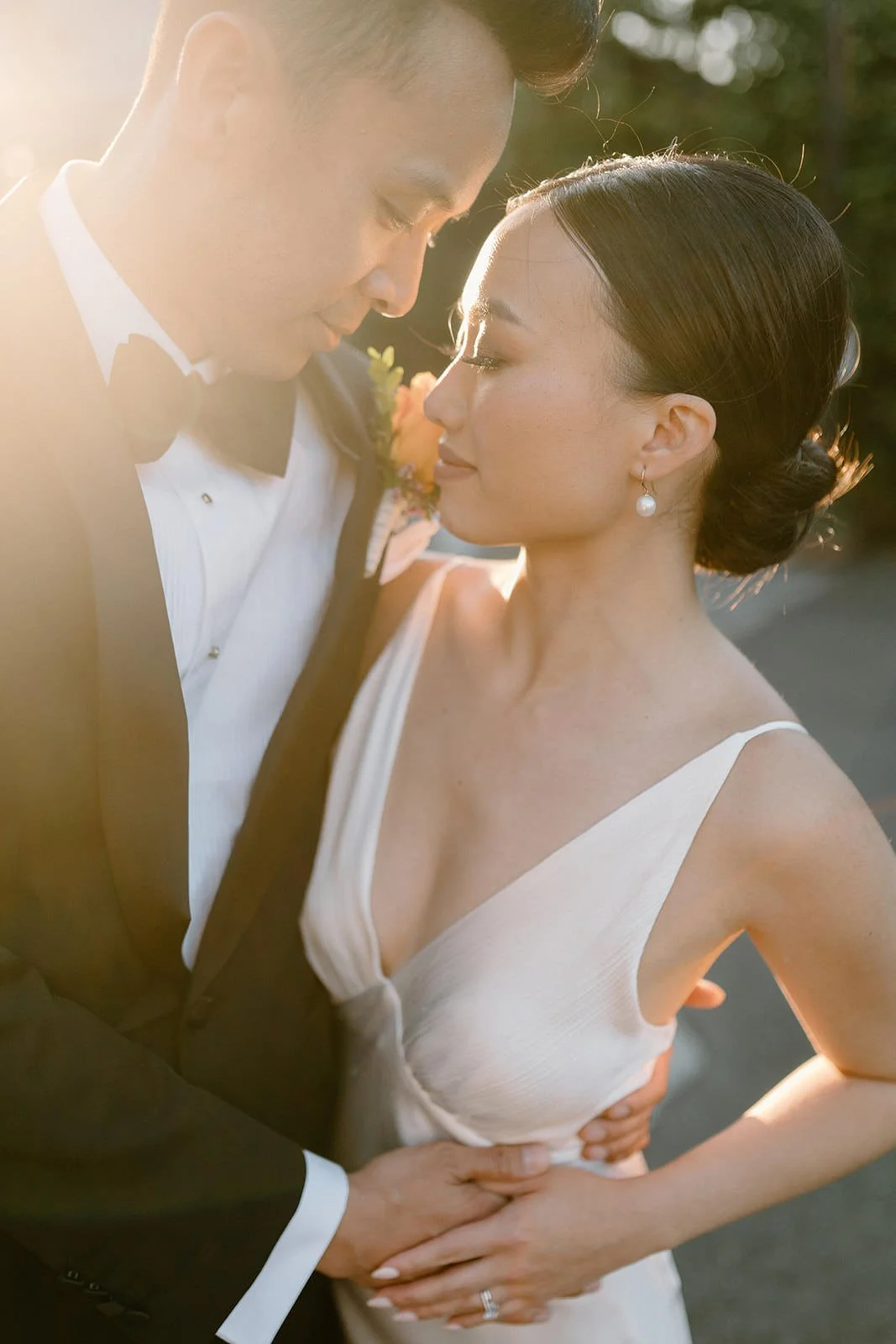 A newlywed couple in wedding attire standing close together, smiling in an intimate moment during sunset with sunlight backlighting them.