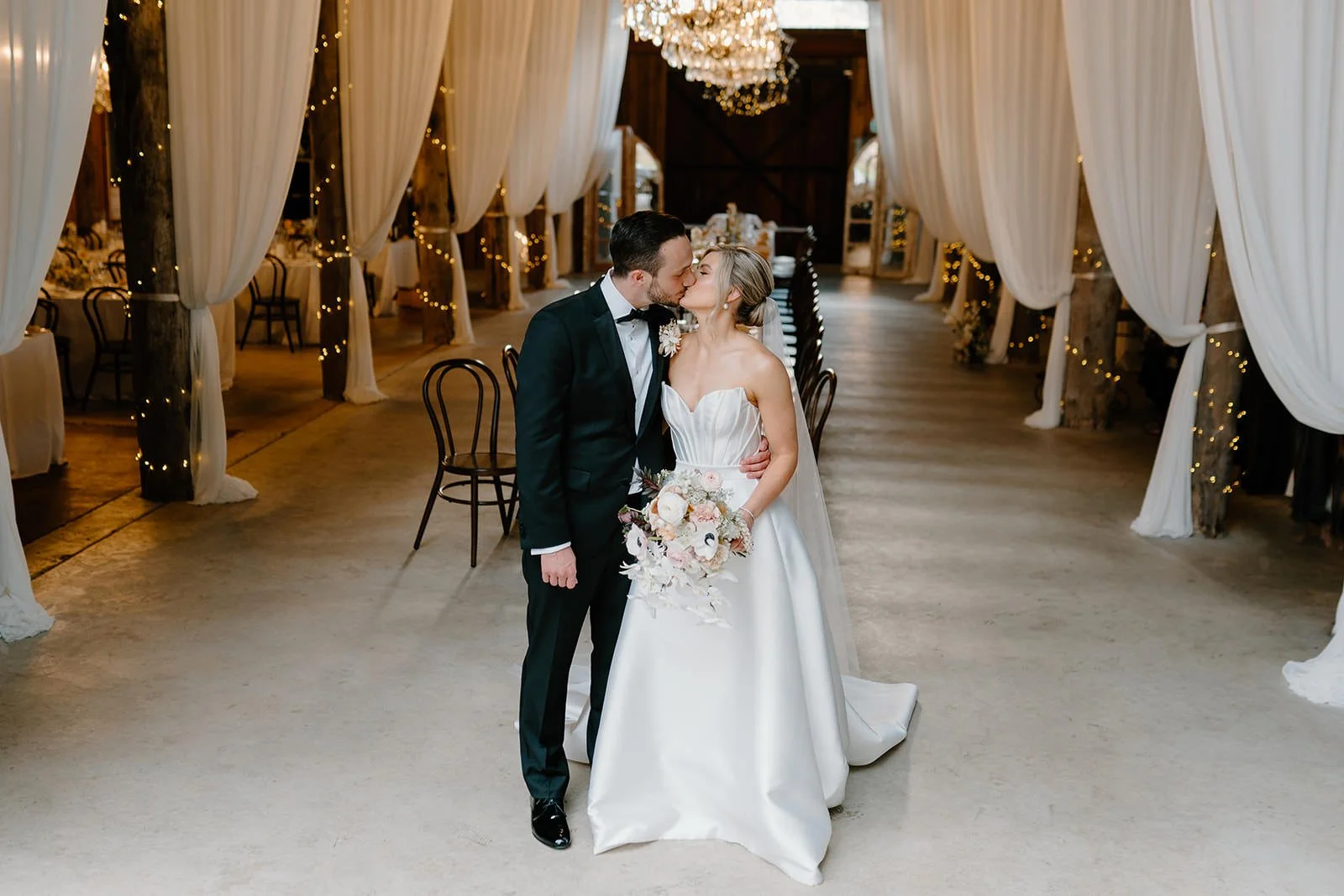 A bride and groom share a kiss at their wedding reception inside a decorated barn venue with draped white curtains and string lights.