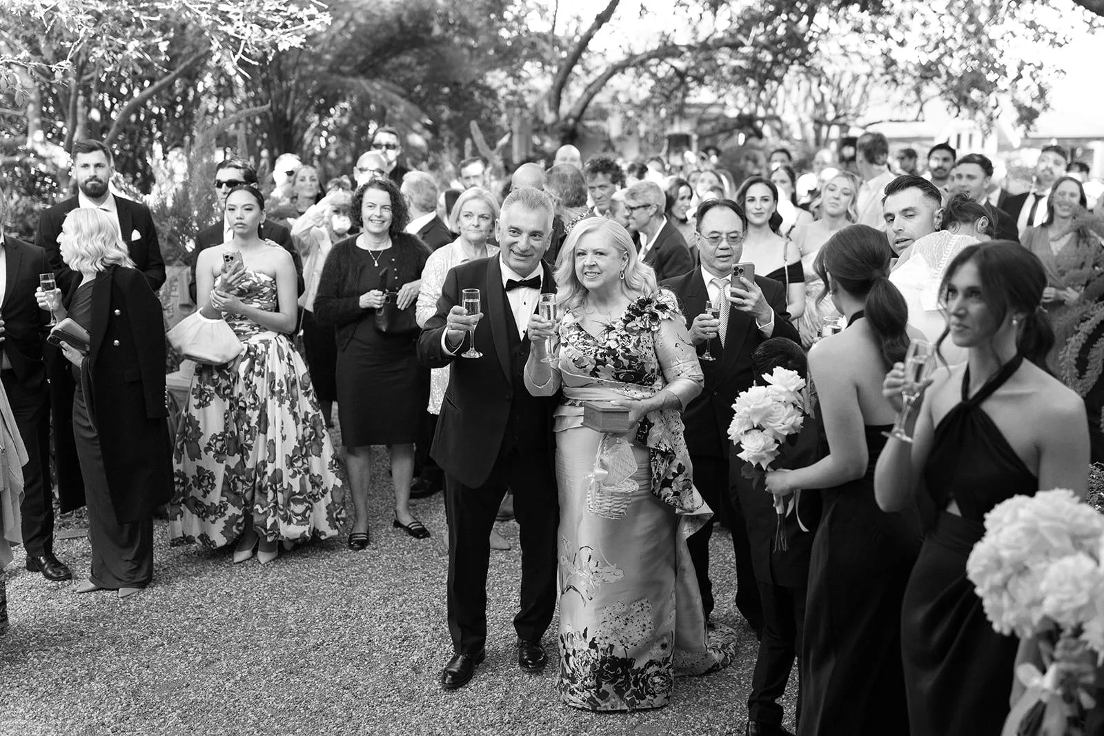 Black and white photo of a crowd of people dressed in formal attire, celebrating outdoors with drinks, some smiling and toasting.