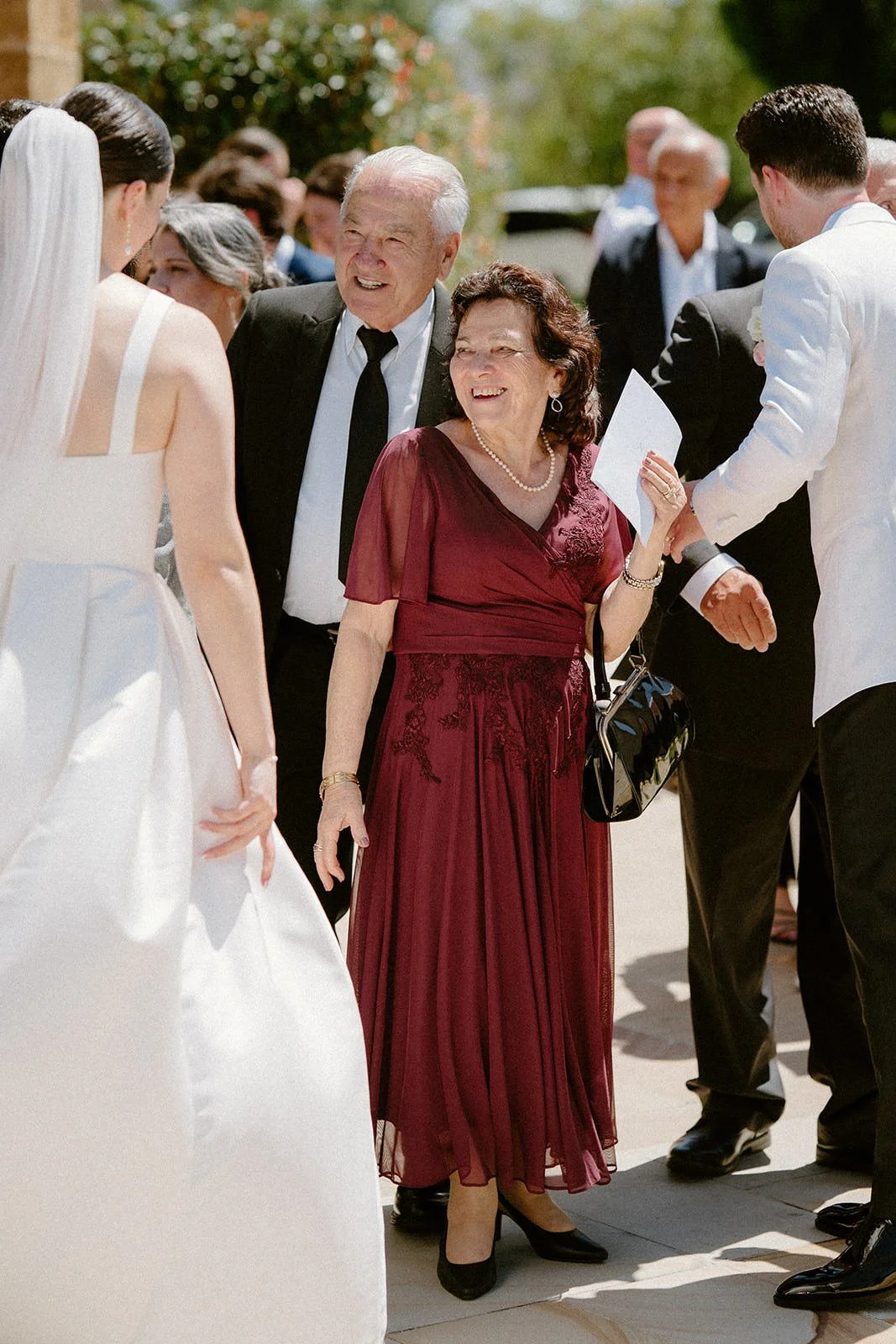 People gathered outdoors at a wedding, with a woman in a burgundy dress smiling and holding an envelope, surrounded by other guests.