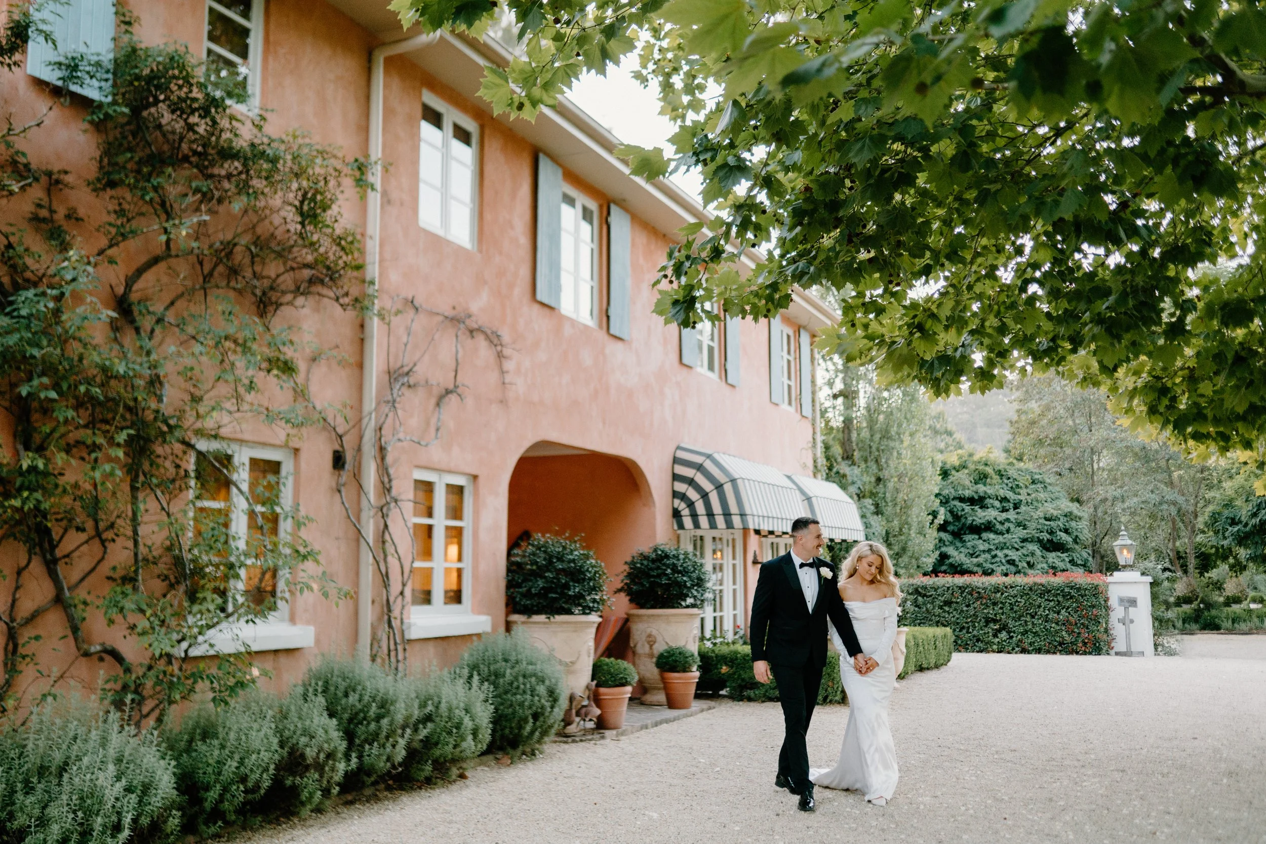 A newlywed couple walking hand in hand outside a pink-colored building with green trees overhead.
