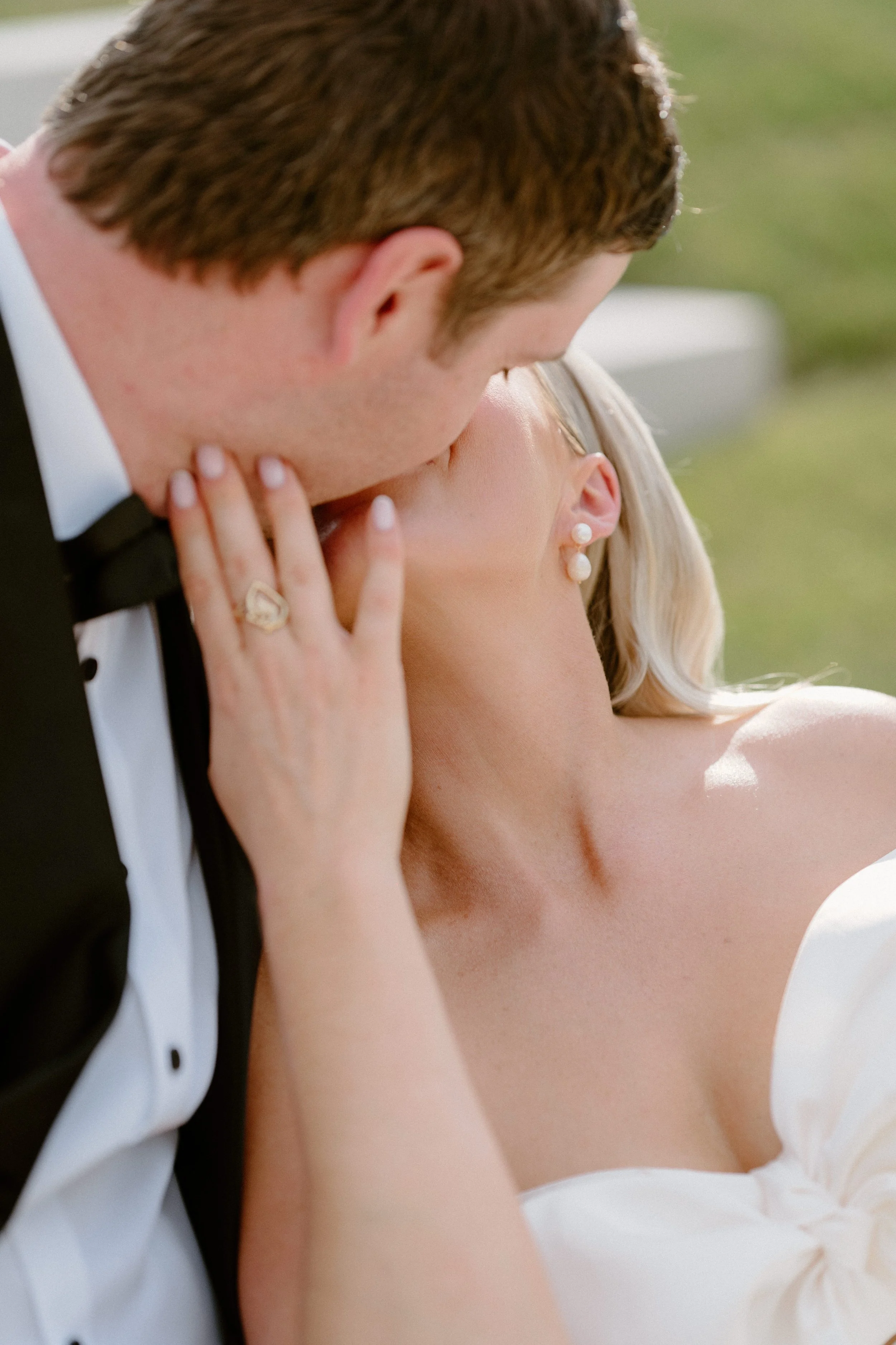 A close-up of a couple kissing, with the man in a tuxedo and the woman wearing pearl earrings, outdoors with greenery in the background. The Sienna wedding photographer.