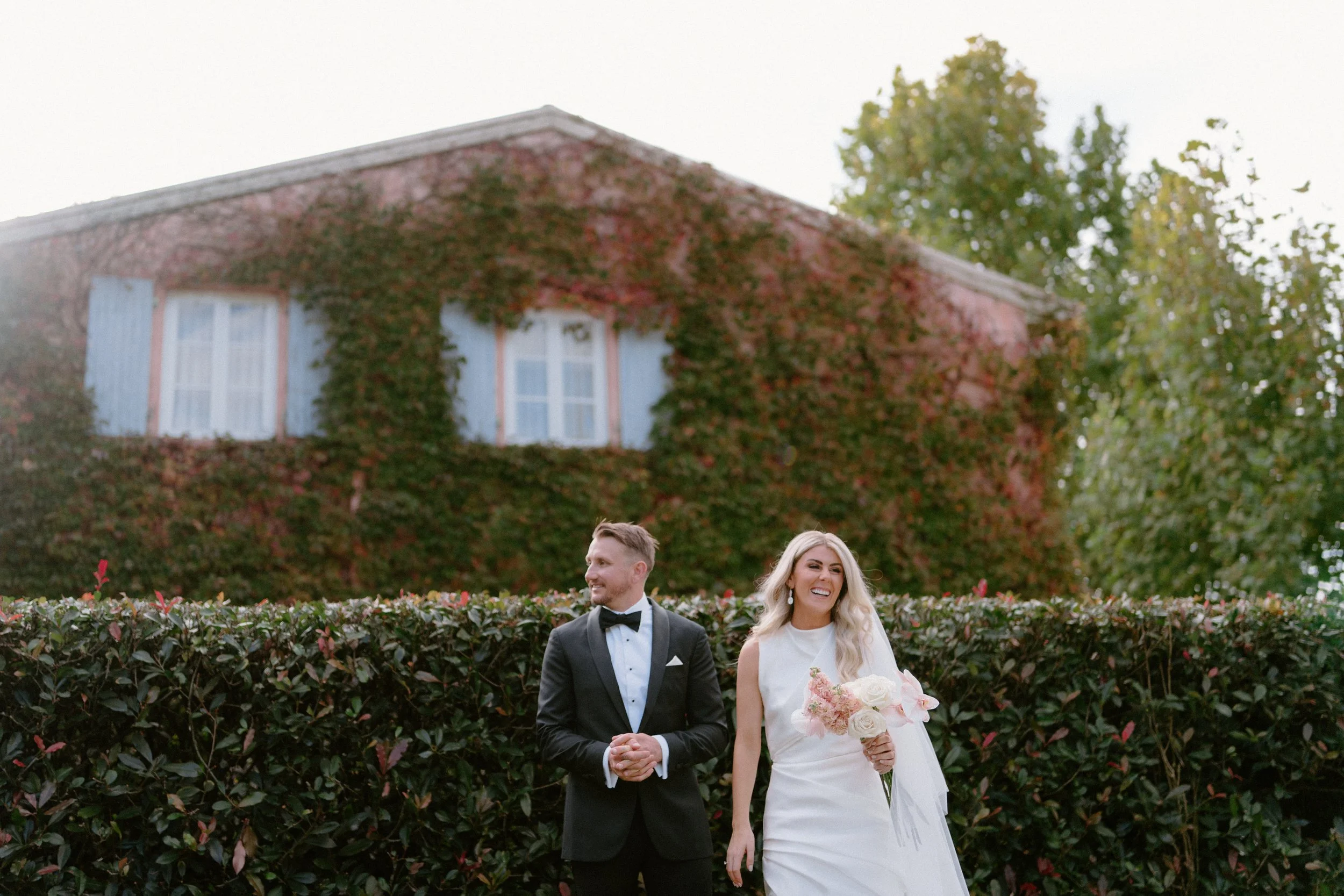 A newlywed couple stands outdoors, the bride holding a bouquet of light pink and white flowers, the groom in a black tuxedo with a bowtie, smiling and looking away, with a background of green hedge and a brick house covered in red and green ivy trees