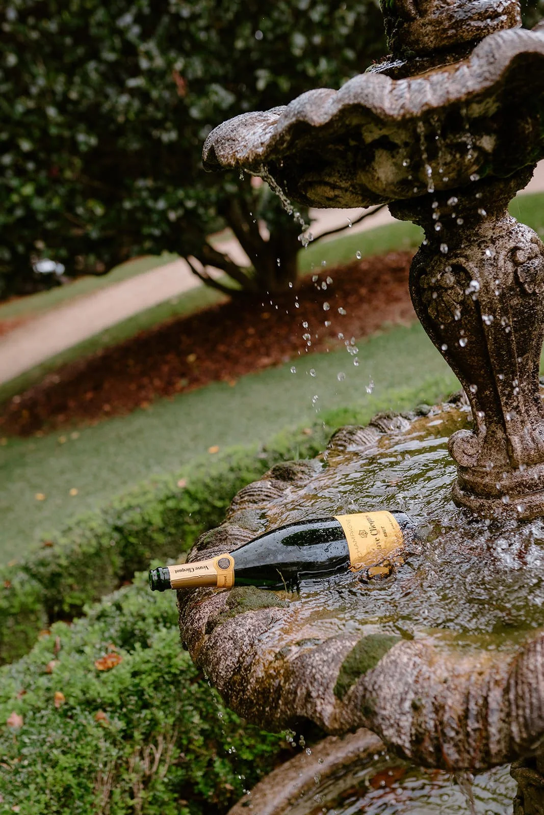 A bottle of champagne resting on the lower tier of a stone fountain with water flowing from the upper tier, set in a landscaped garden.