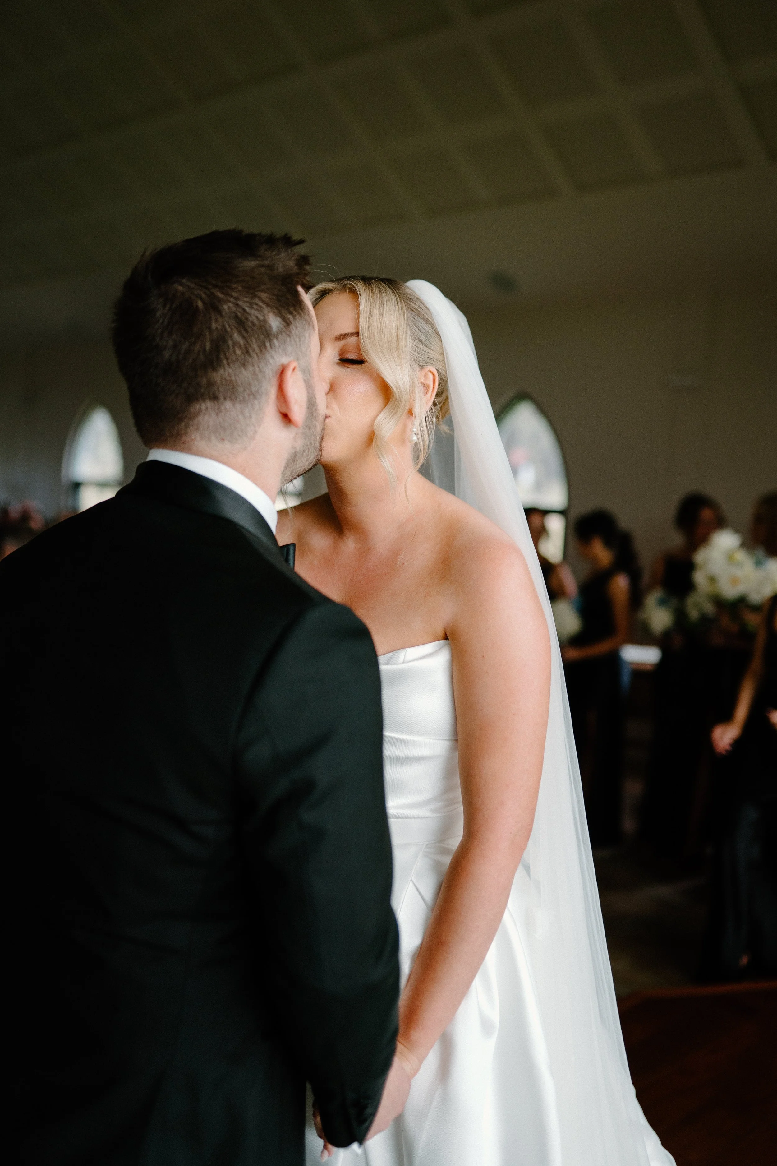 A bride and groom kiss during their wedding ceremony. The bride wears a strapless white gown and veil, while the groom is in a black tuxedo. Guests are visible in the background.