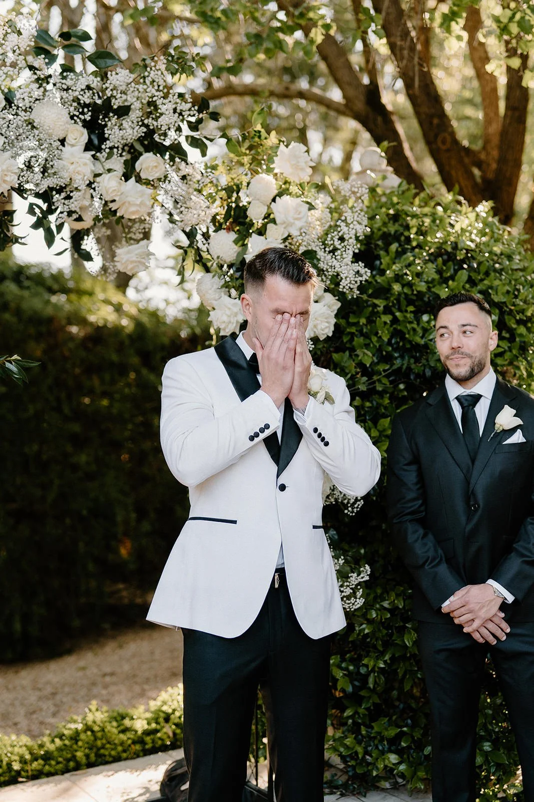 Groom in a white tuxedo with black accents covering his face, standing under a floral wedding arch next to another man in a black tuxedo, outdoors.