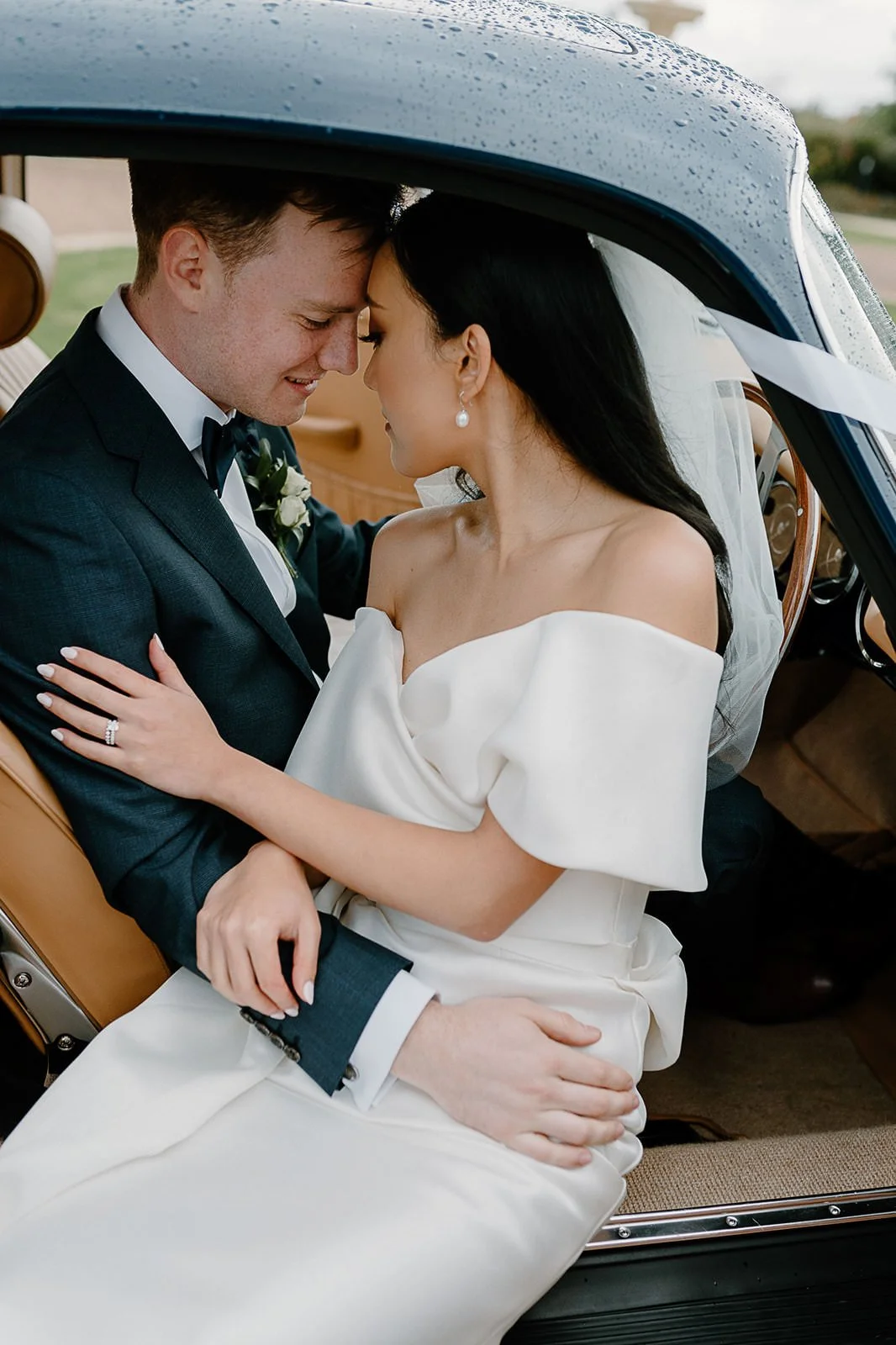 A bride and groom sit close together inside a vintage car, touching foreheads and smiling, after their wedding ceremony.