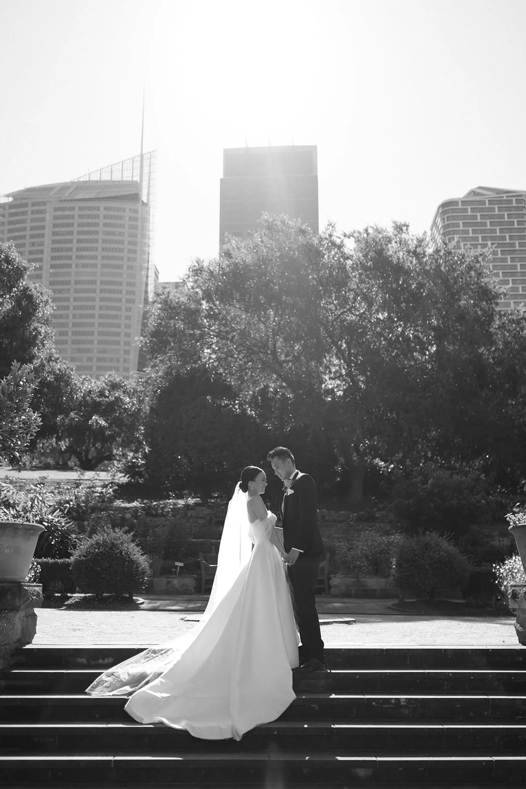 A bride and groom holding hands on stairs outdoors with trees and tall buildings in the background, black and white photograph.