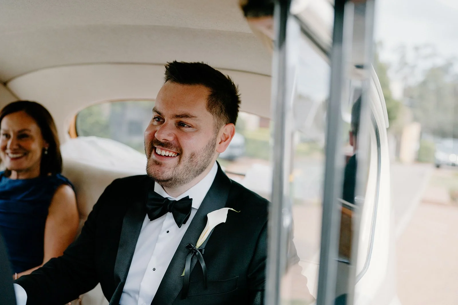 A smiling man in a tuxedo with a bow tie and a white calla lily boutonniere sits inside a car with a woman in a blue dress, both appearing happy and dressed for a wedding or formal event.