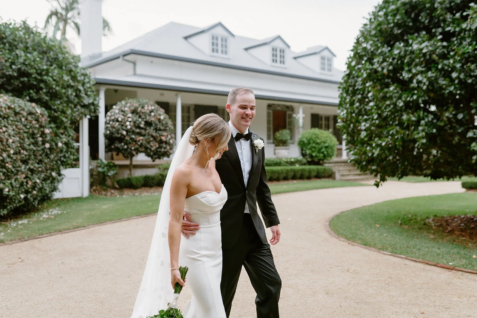 A bride and groom walking together outdoors near a white house with greenery in the background.