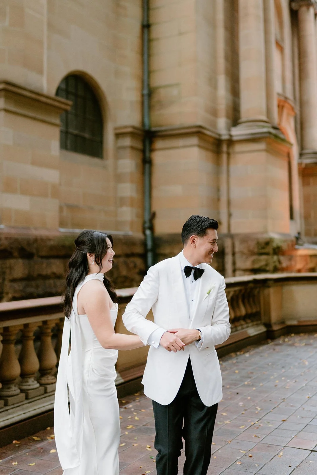 A newlywed couple holding hands and smiling outside a historic building with stone walls and arched windows during their wedding ceremony.