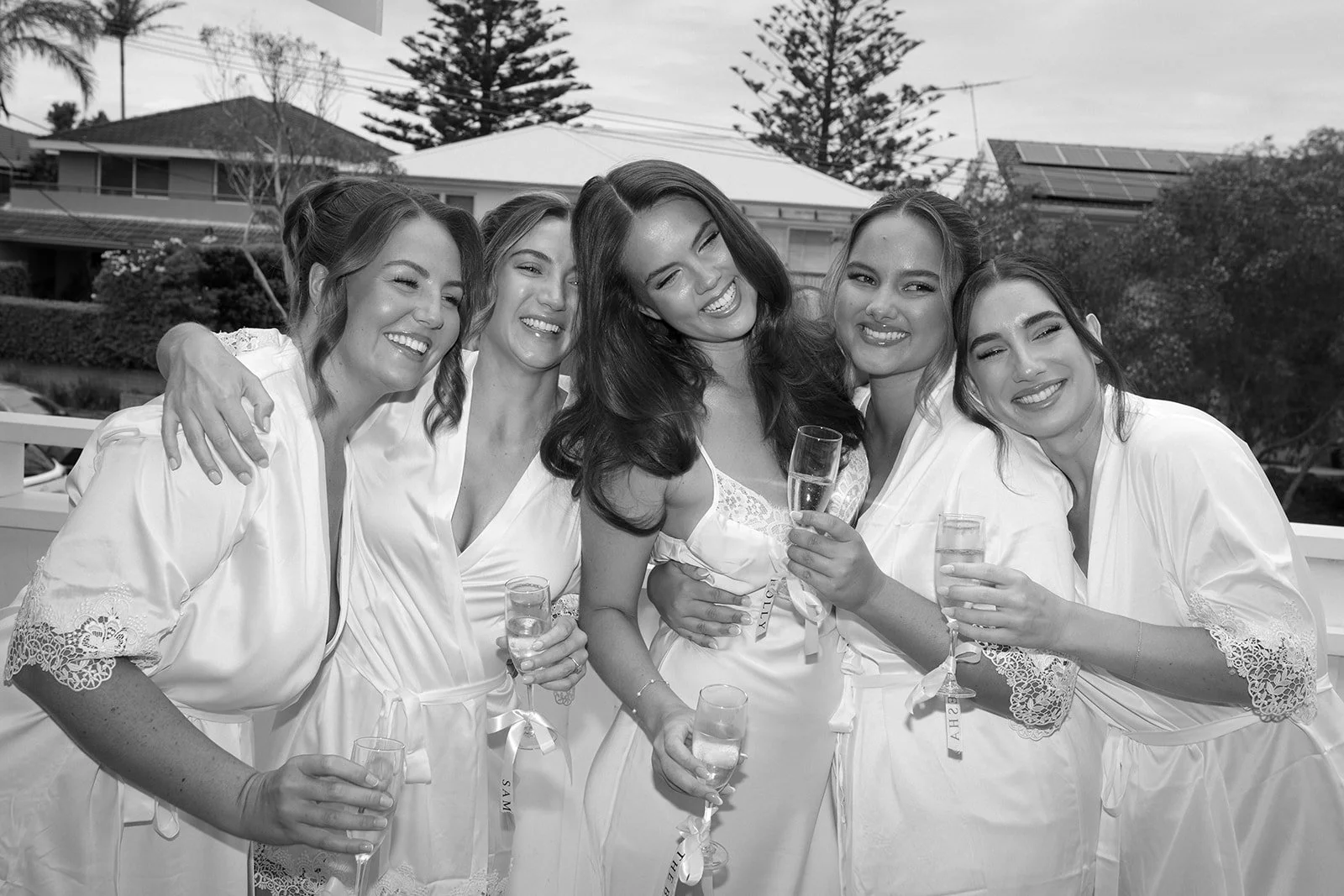 Five women in matching robes smiling and holding glasses of champagne during a bridal celebration outdoors.