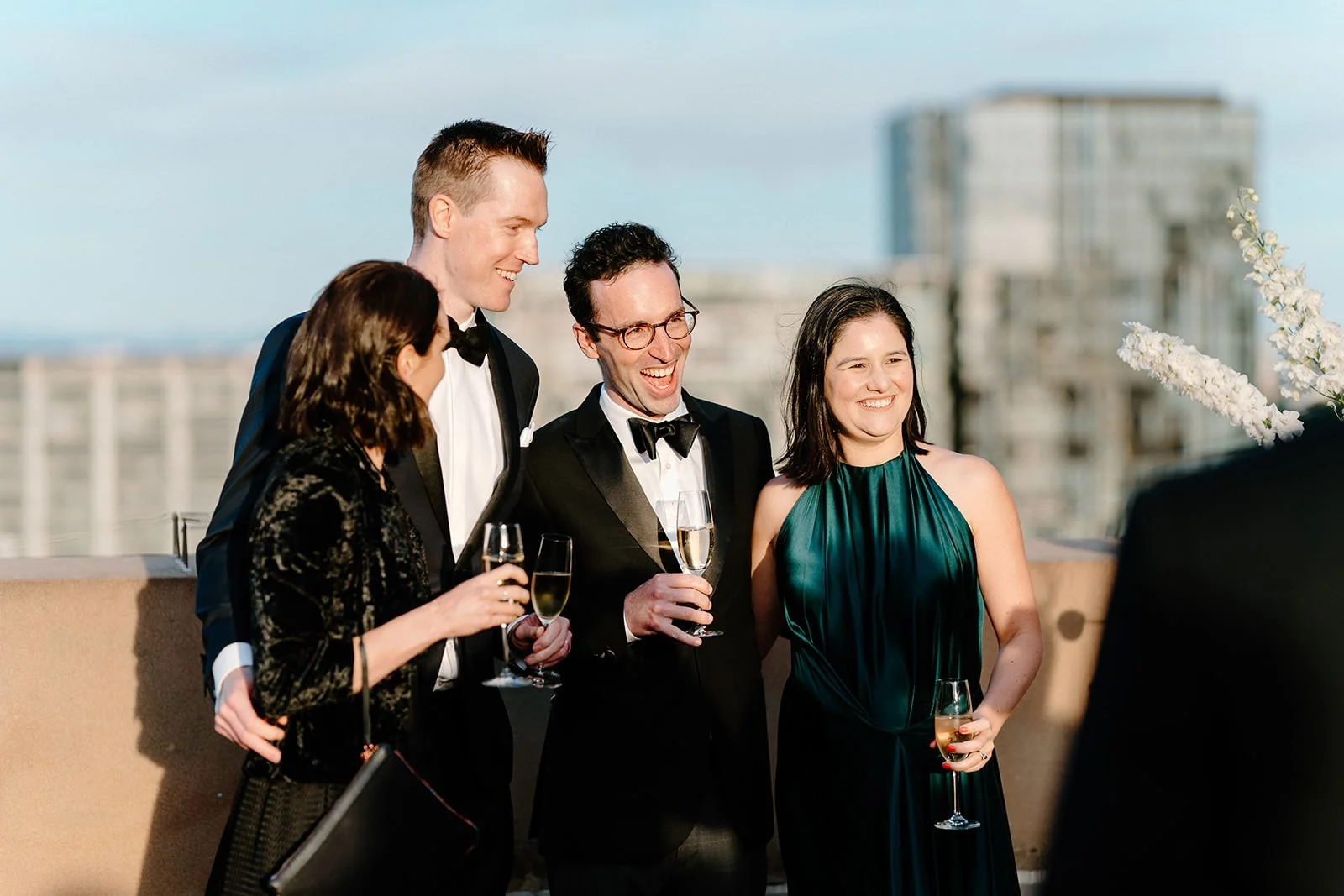 Group of five people celebrating at a rooftop event, dressed in formal attire, holding champagne glasses, smiling and enjoying the moment with city buildings in the background.