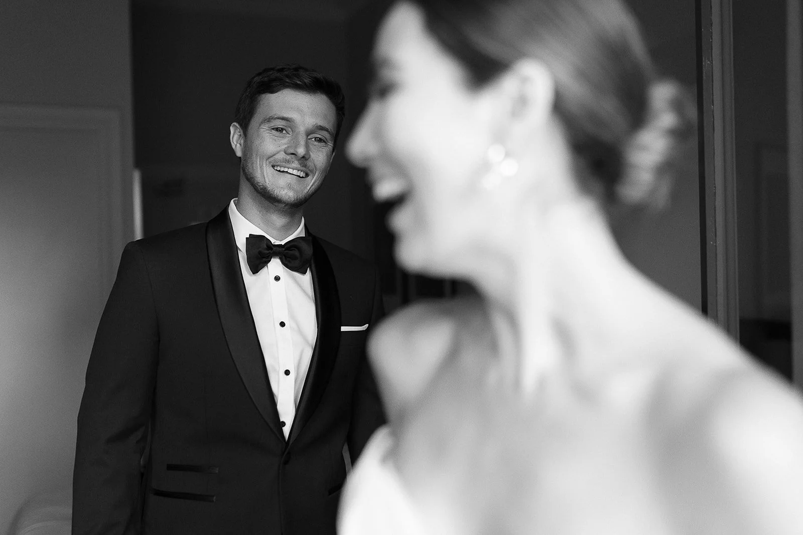 A smiling man in a tuxedo looking at a woman in a wedding dress, with the woman mostly out of focus in the foreground.