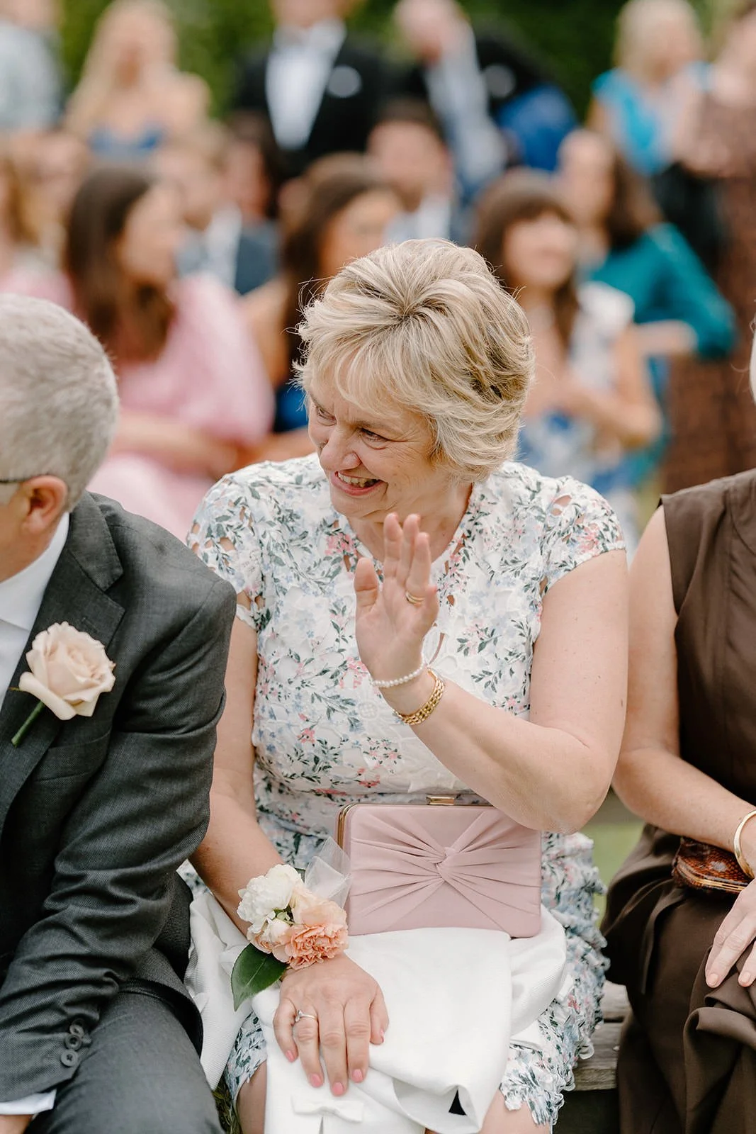 A woman with short blonde hair smiling and laughing at a wedding or event, sitting next to a man in a suit with a boutonniere, in front of a group of seated guests.