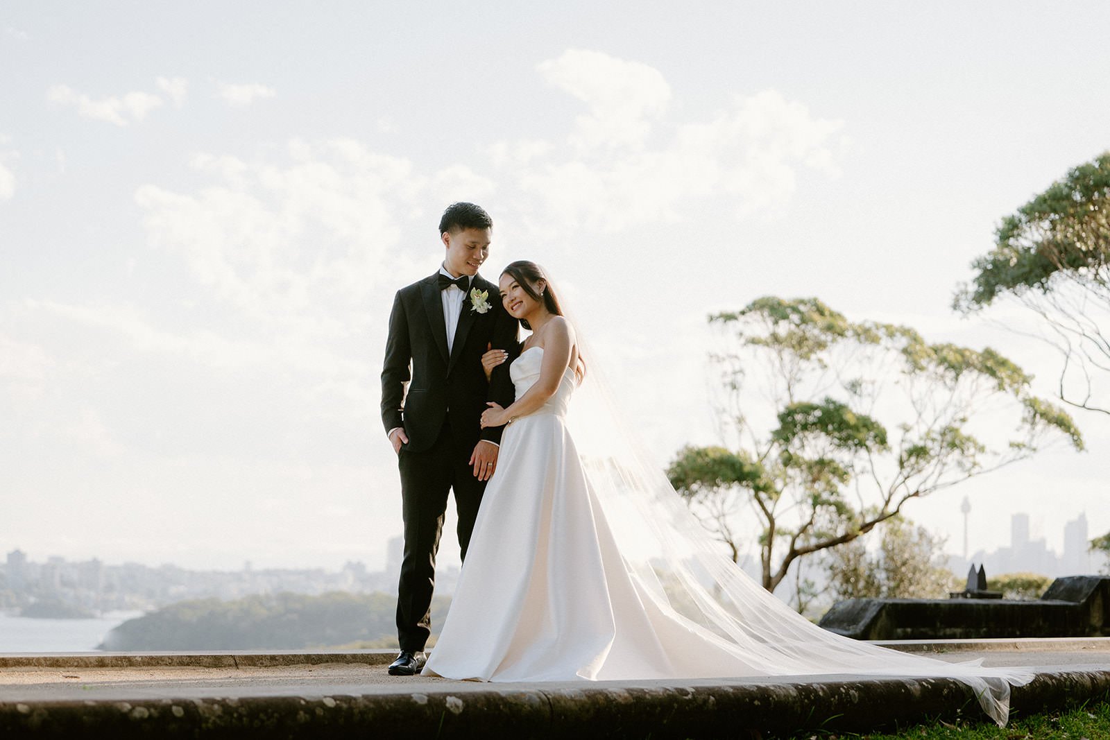 A bride and groom standing outdoors on a platform with city skyline in the background, dressed in wedding attire. The groom is in a black tuxedo and the bride in a strapless white wedding gown with a long train.