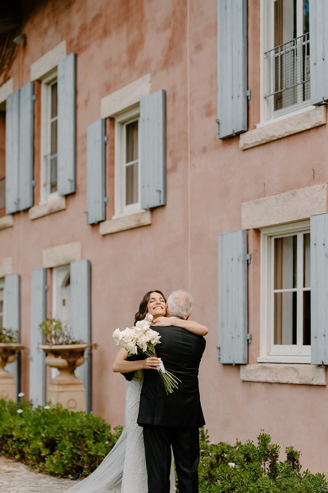 A bride in a white dress hugging an elderly man in a black suit outside a pink building with blue window shutters, holding a bouquet of white flowers.