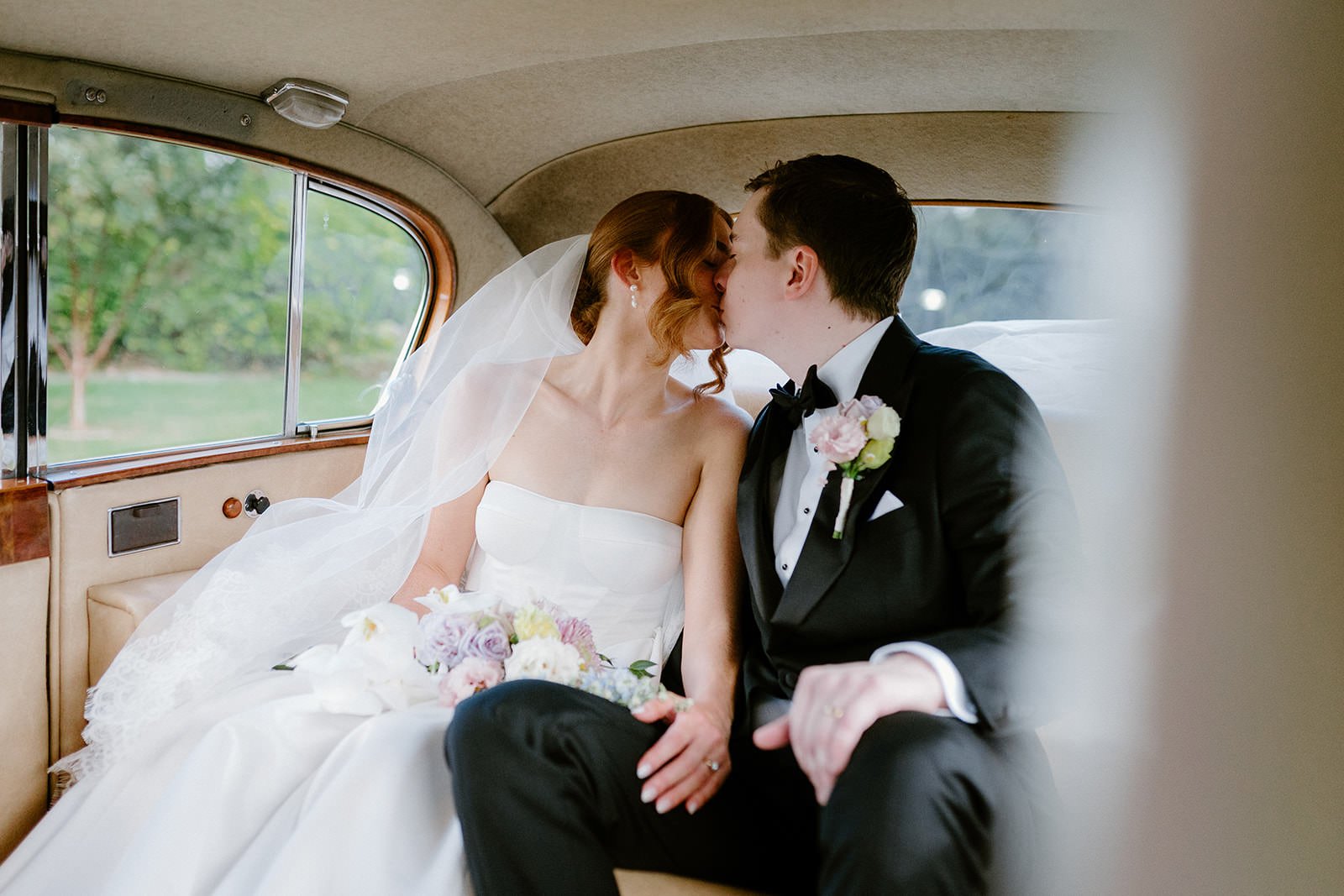 Bride and groom kissing inside a vintage car on their wedding day, with greenery visible outside the windows.